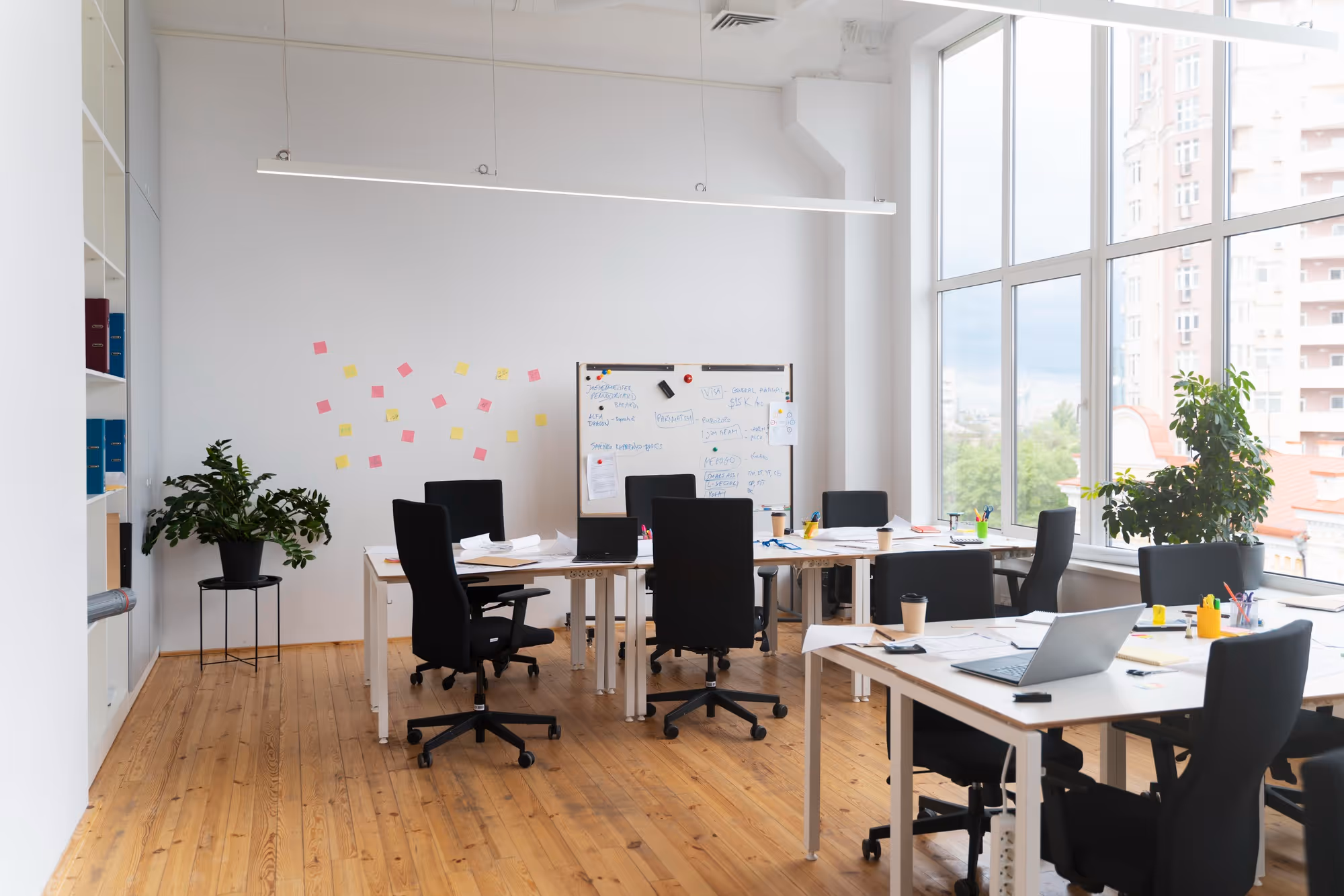 Bright modern office with large windows, several black chairs around white desks, laptops, papers, plants, and a whiteboard with notes and sticky notes on the wall.