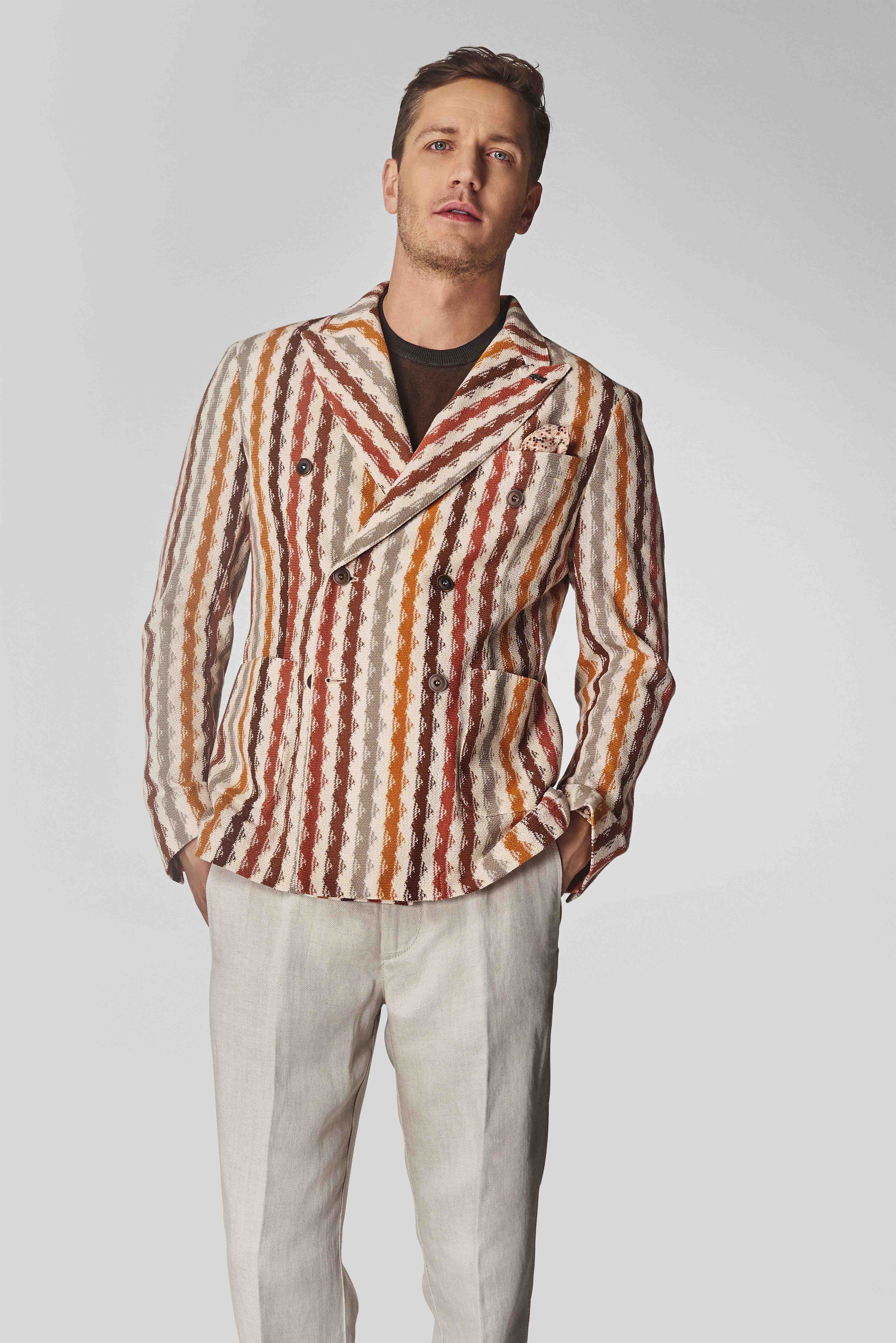 Mann trägt einen zweireihigen Blazer mit bunten Streifen in Rot-, Orange- und Erdtones sowie helle Hose vor grauem Hintergrund.