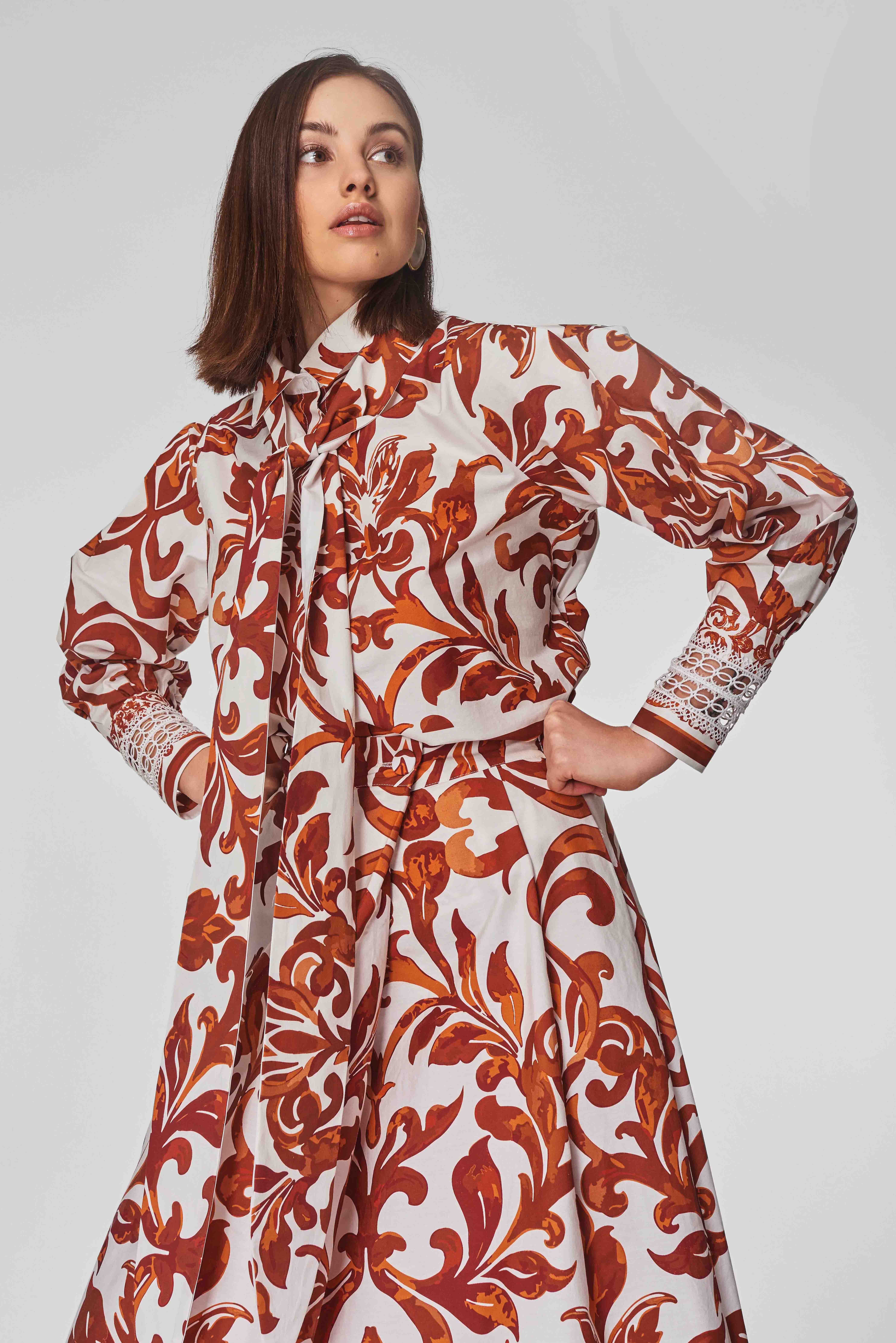 Frau in einem langärmeligen Kleid mit orange-rotem Blumenmuster und weißer Spitzenmanschette posiert vor grauem Hintergrund.