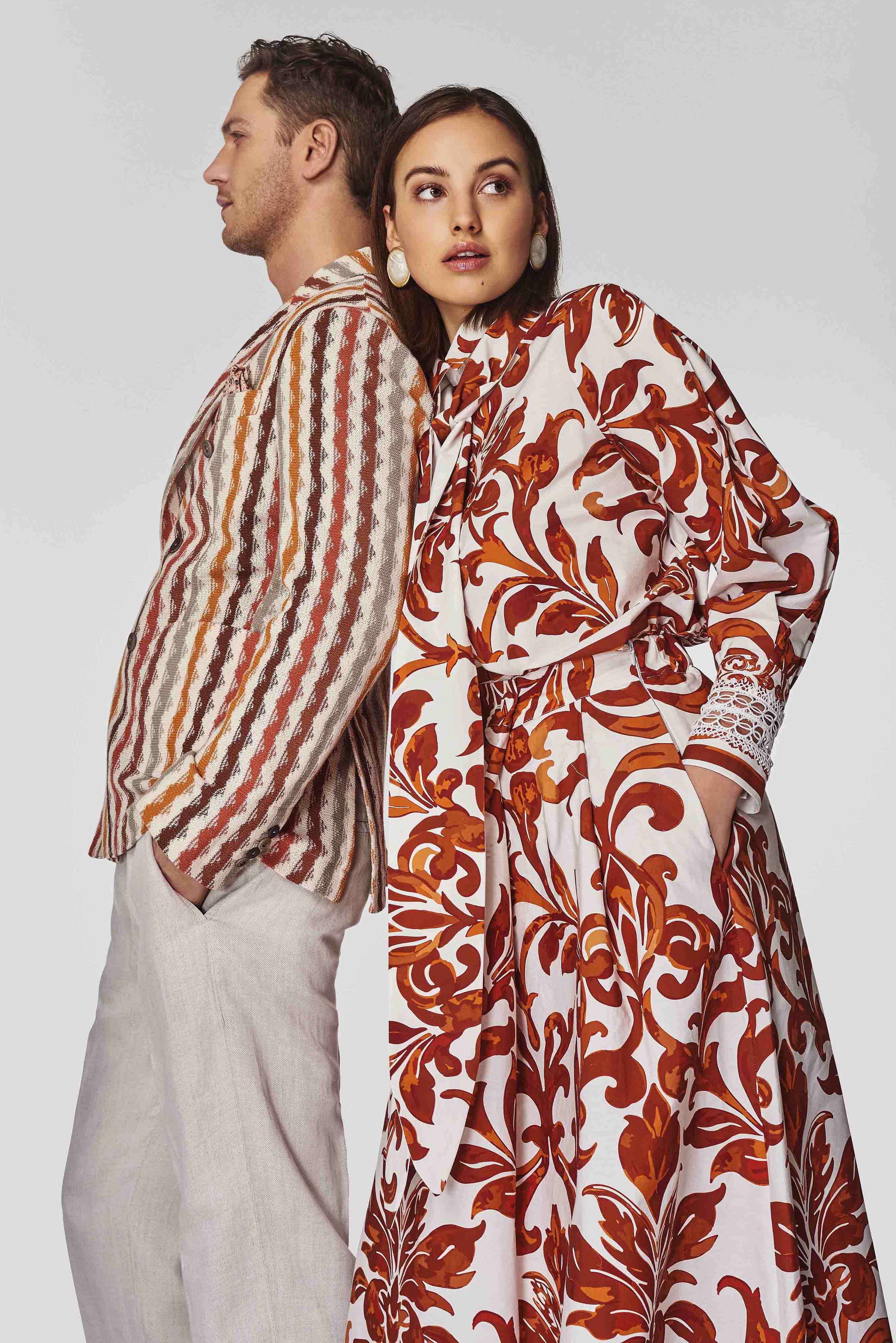 Ein Frau und ein Mann lehnen Rücken an Rücken; die Frau trägt ein weißes Kleid mit roten und orangefarbenen floralen Mustern, der Mann eine gestreifte Jacke in Erdtönen und beige Hosen.