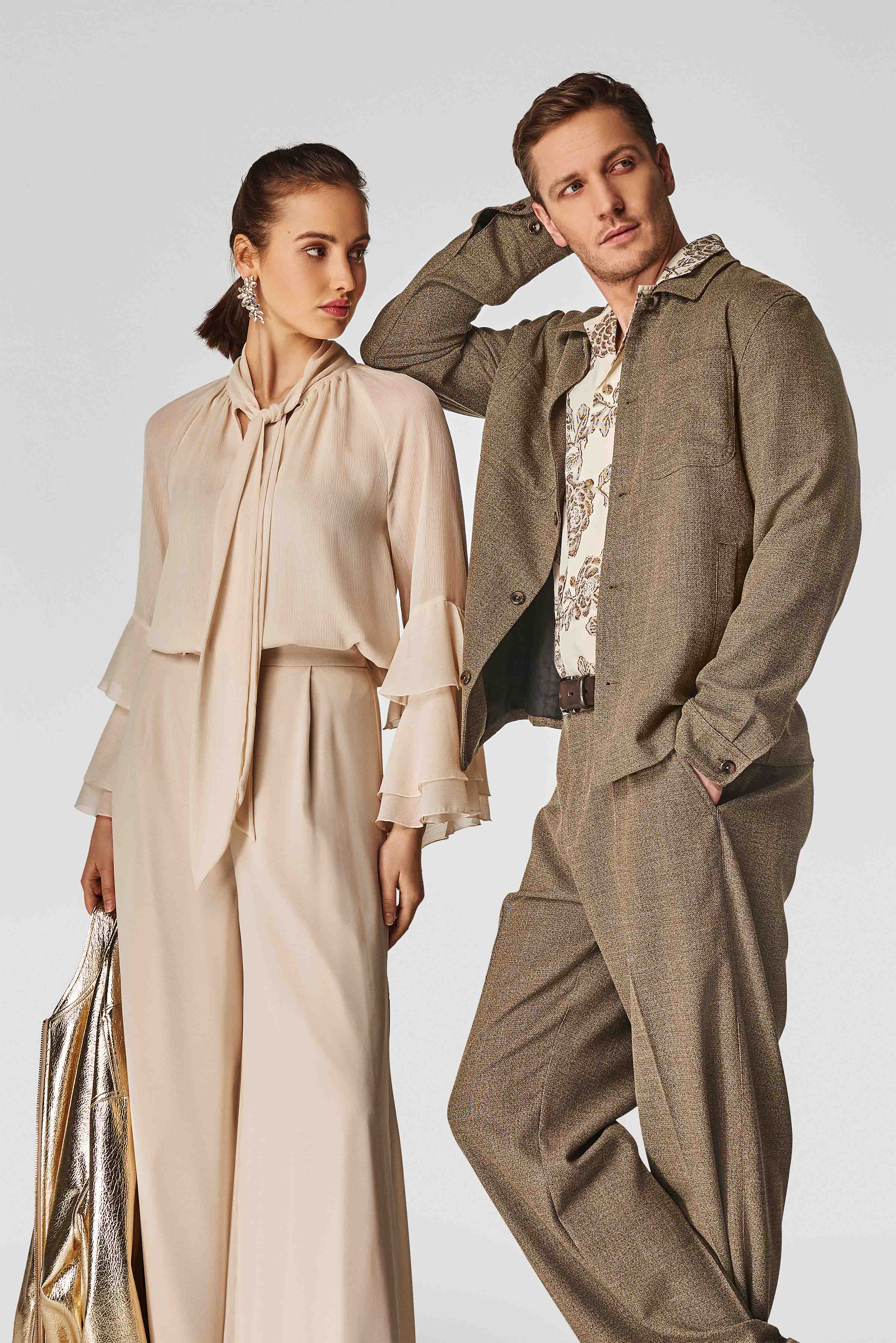 Frau in beiger Bluse mit Volantärmeln und weitem Hosenrock hält eine goldene Tasche, neben einem Mann in beigem Anzug und floralem Hemd.