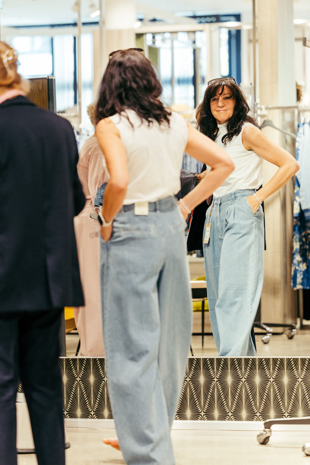 Frau mit weißen Ärmellosem Oberteil und weiten blauen Jeans schaut in einem Laden in einen Spiegel.