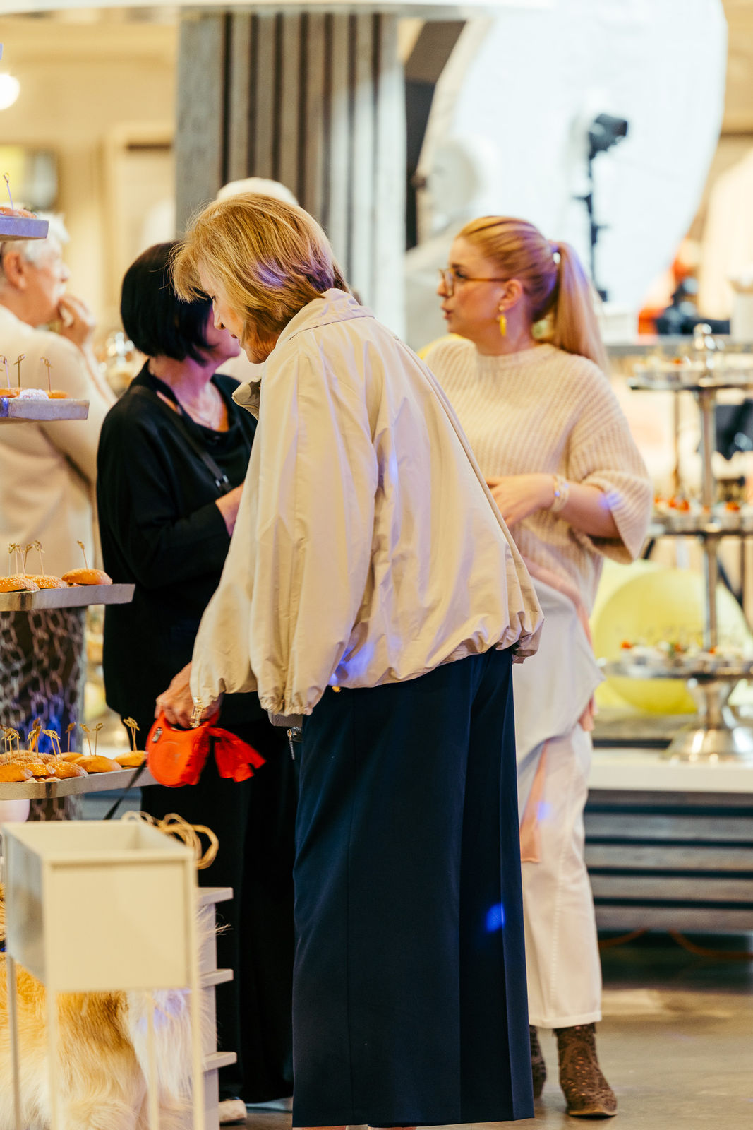 Ältere Frau mit Hund an der Leine greift nach kleinen belegten Brötchen an einem Buffet, während andere Frauen im Hintergrund sprechen.