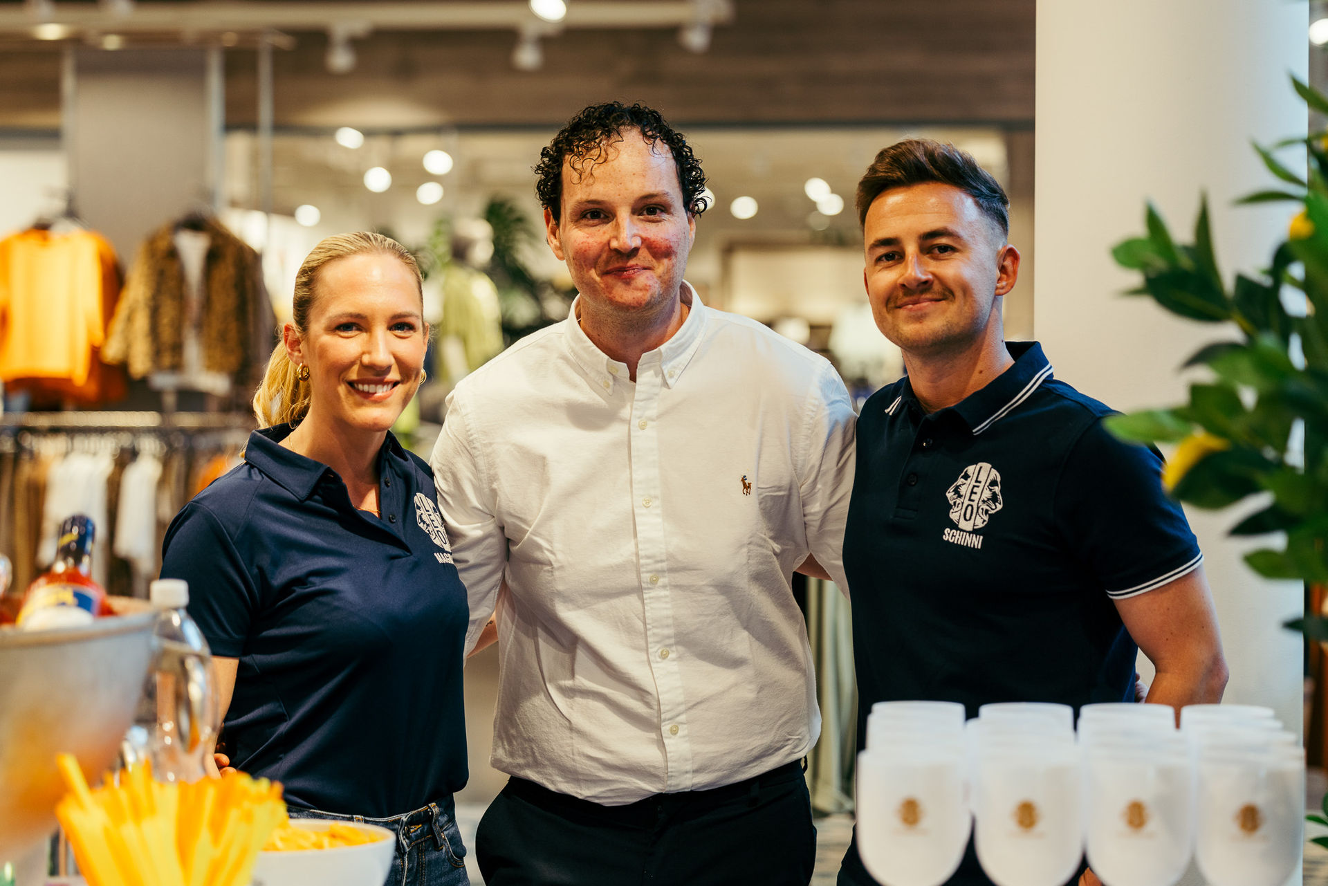 Drei lächelnde Personen in einem Geschäft, zwei tragen dunkelblaue Poloshirts mit Logos, eine Person trägt ein weißes Hemd.