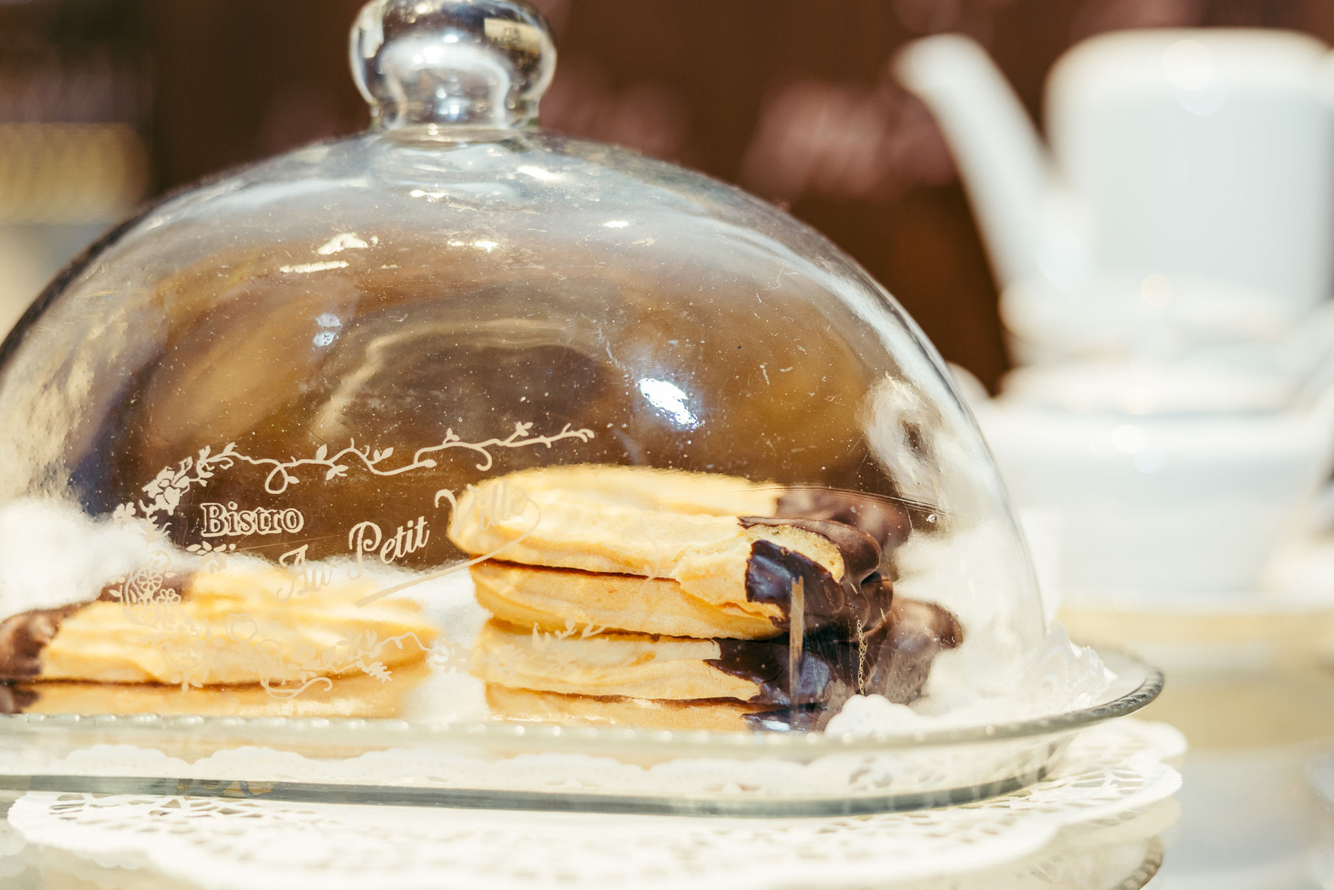 Glasglocke mit Schokoladendip-Plätzchen auf weißer Spitze im Bistro-Setting mit Teekanne im Hintergrund.