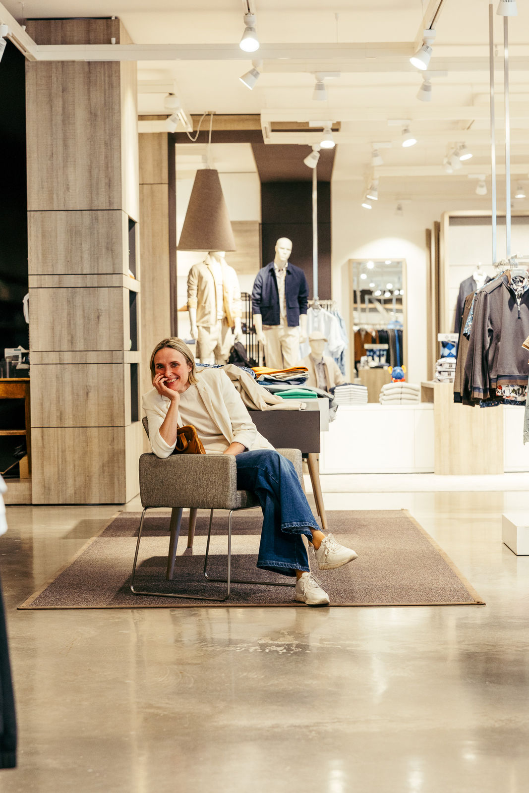 Frau in weißem Oberteil und blauer Jeans sitzt lächelnd auf einem Stuhl in einem Bekleidungsgeschäft.