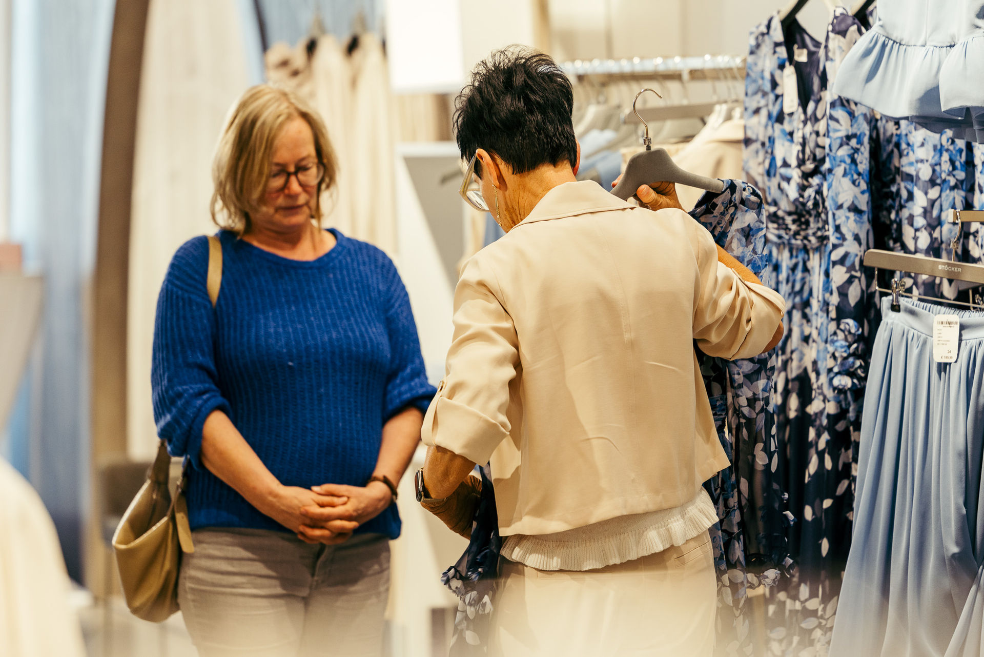 Zwei Frauen beim Einkaufen, eine hält ein blau gemustertes Kleid in der Hand, die andere schaut zu und trägt einen blauen Pullover und eine Tasche.