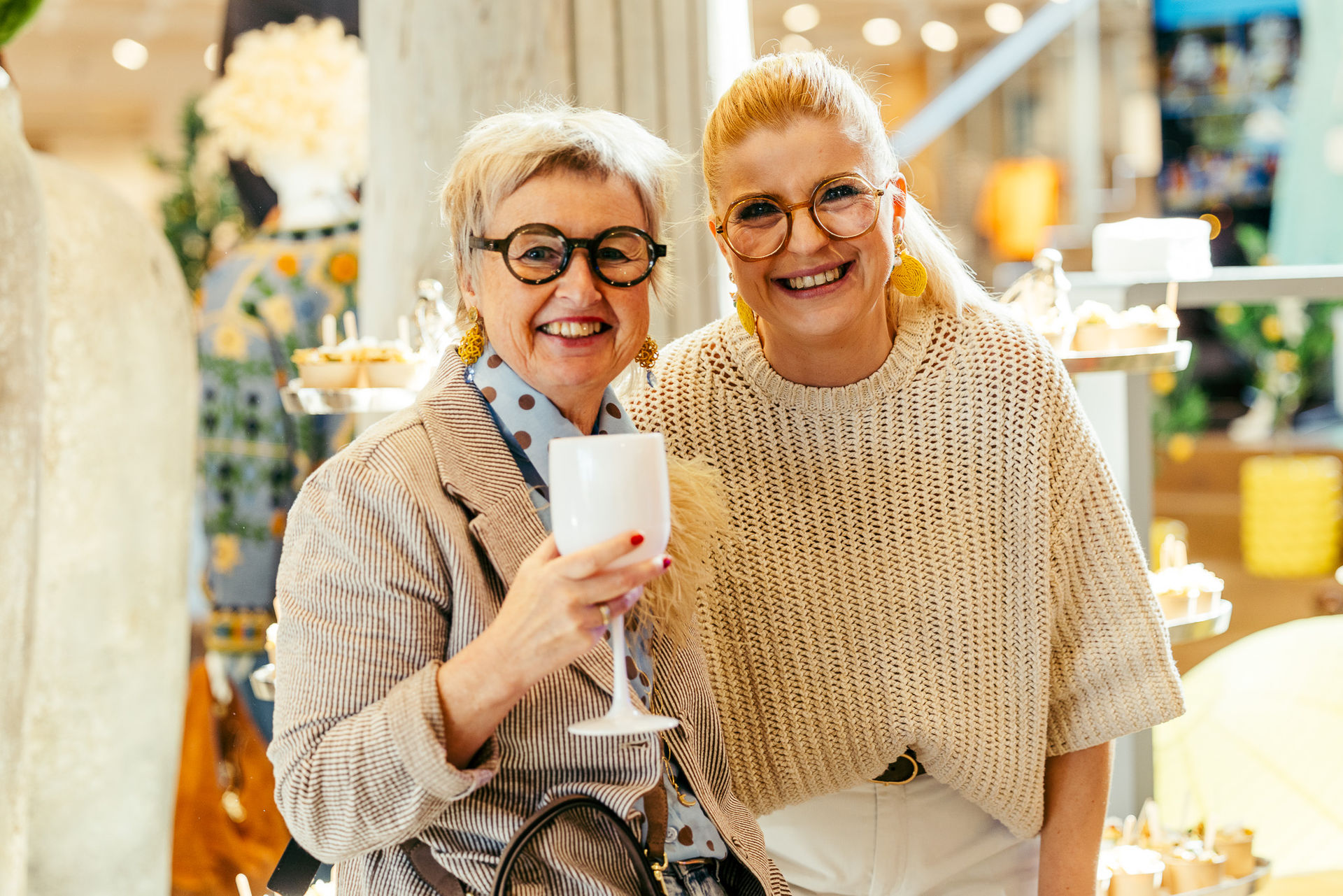 Zwei lächelnde Frauen mit Brillen in einem hellen Raum, eine hält ein Glas, im Hintergrund Kuchen und Dekoration.