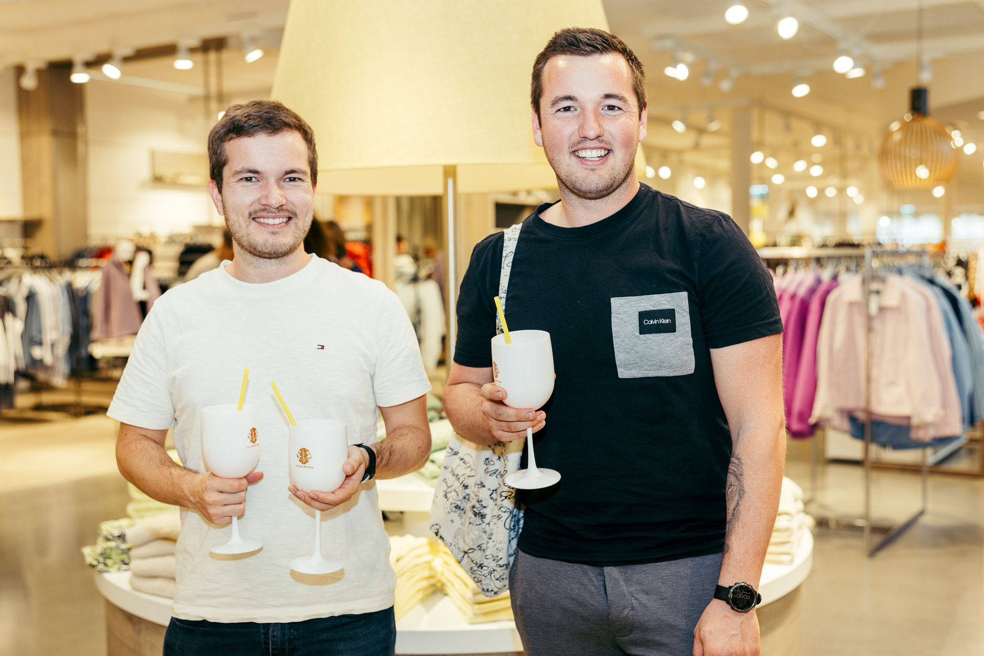 Zwei lächelnde junge Männer halten jeweils ein weißes Glas mit gelbem Strohhalm in einem Bekleidungsgeschäft.