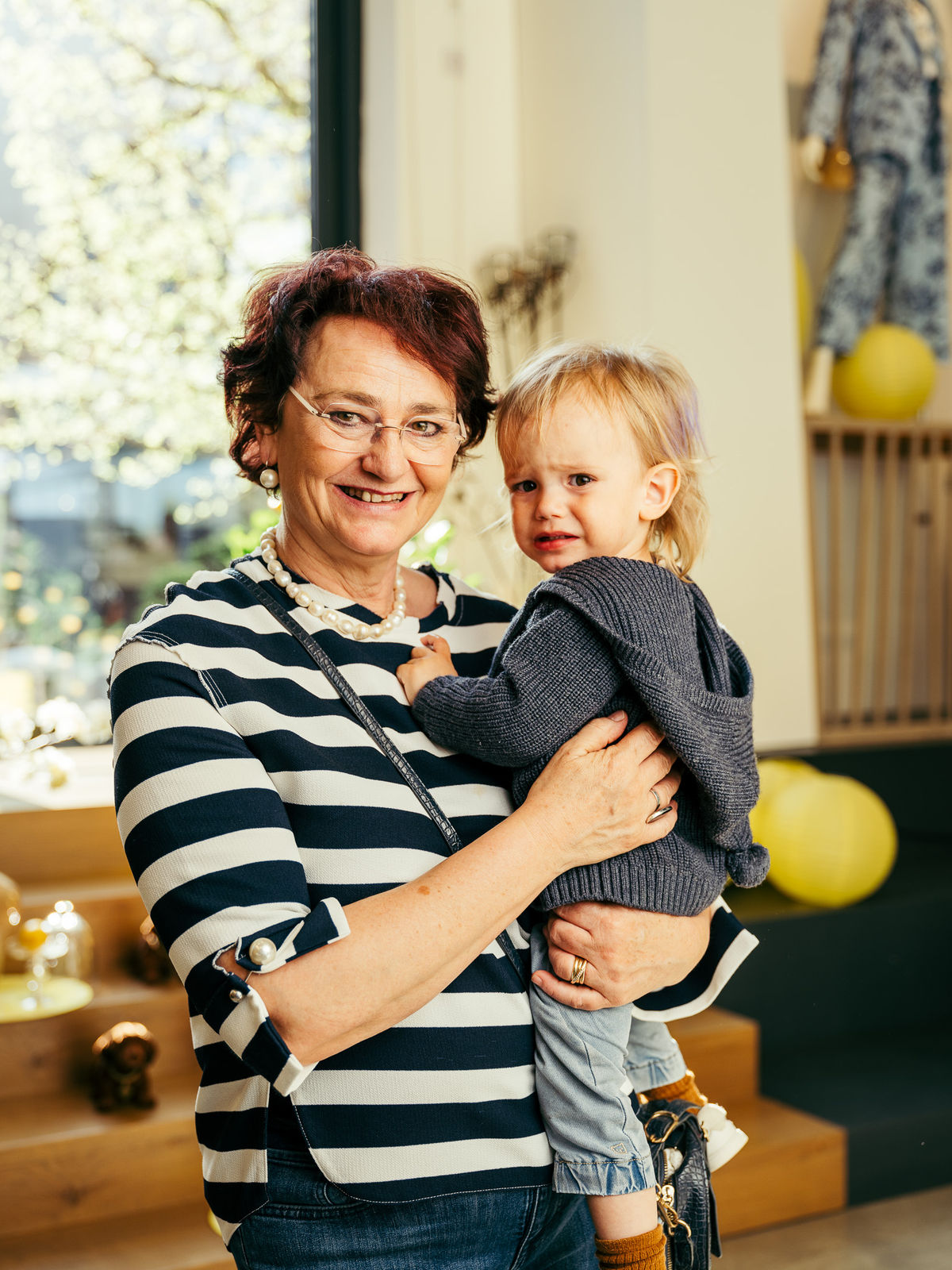 Ältere Frau mit Brille und gestreiftem Pullover hält ein sorgenvoll dreinblickendes Kleinkind in einem hellen Innenraum.