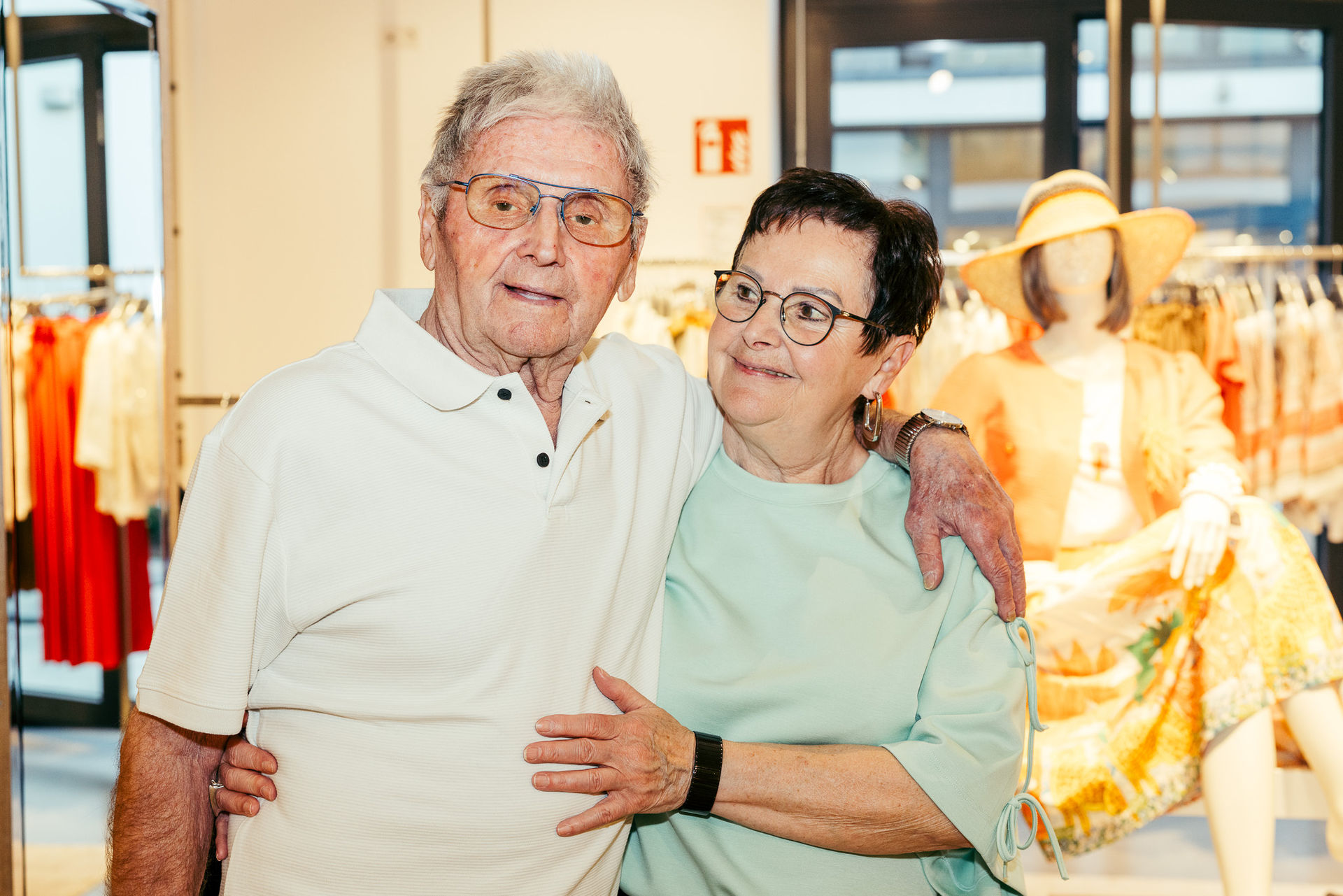 Älteres Paar umarmt sich herzlich in einem Bekleidungsgeschäft mit einer Schaufensterpuppe im Hintergrund.