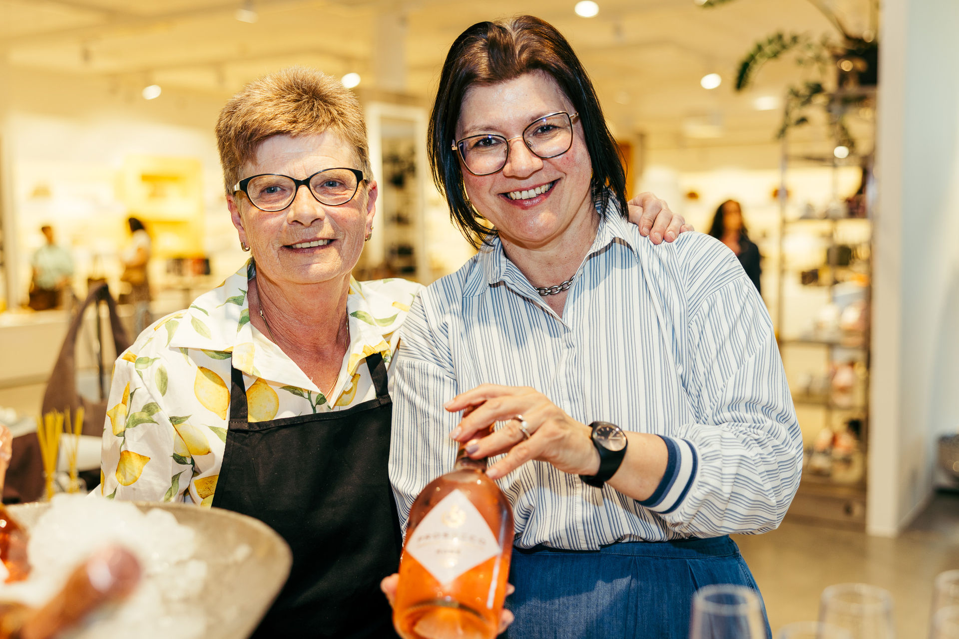 Zwei lächelnde Frauen mit Brillen, eine trägt ein schwarzes Latzschürze und die andere hält eine Flasche Roséwein in einem hell beleuchteten Geschäft.