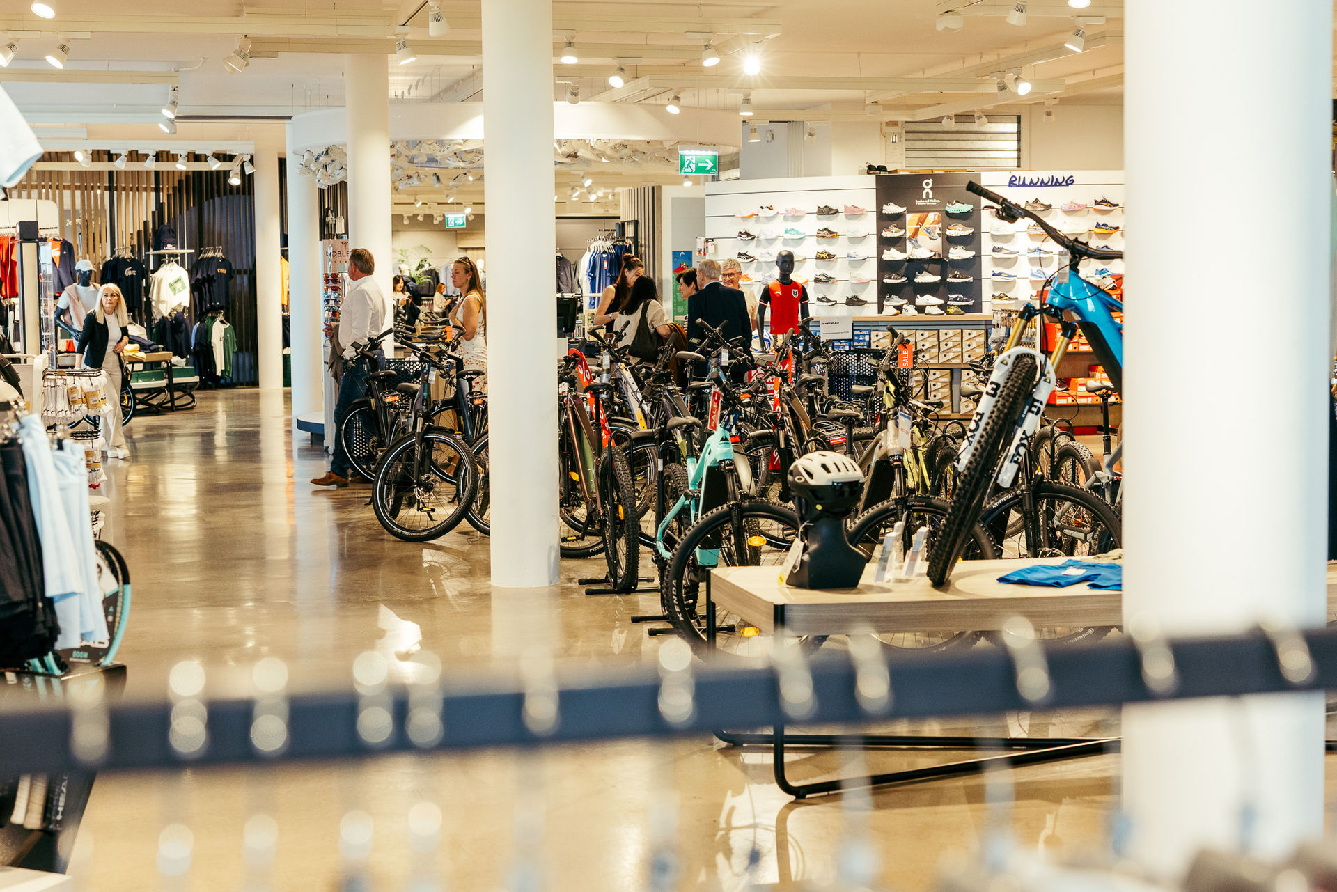 Innenansicht eines gut beleuchteten Sportgeschäfts mit mehreren Fahrrädern, Fahrradhelmen, Sportschuhen an der Wand und Personen beim Einkaufen.
