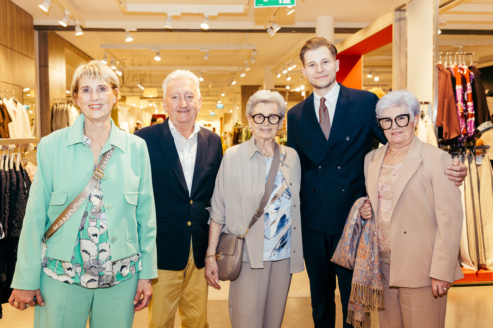 Fünf gut gekleidete Personen in einem Bekleidungsgeschäft, drei ältere Frauen, ein älterer Mann und ein junger Mann im blauen Anzug, lächelnd in die Kamera.