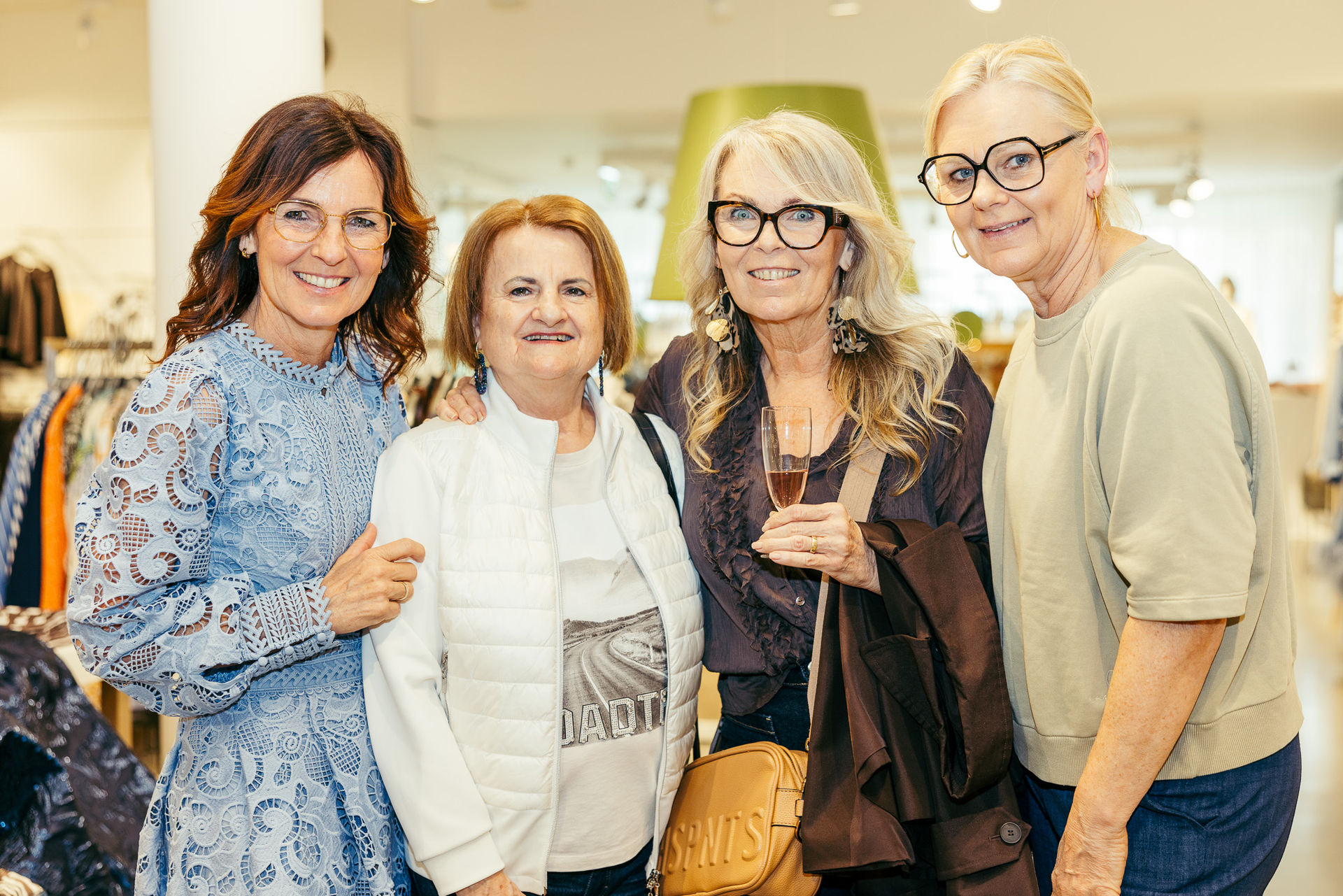 Vier lächelnde Frauen mittleren Alters in einem hellen Raum, eine hält ein Glas mit Getränk.