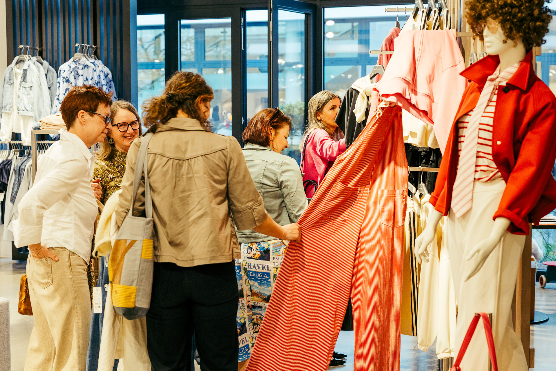 Fünf Frauen schauen in einem Bekleidungsgeschäft auf ein rotes Kleidungsstück und unterhalten sich.