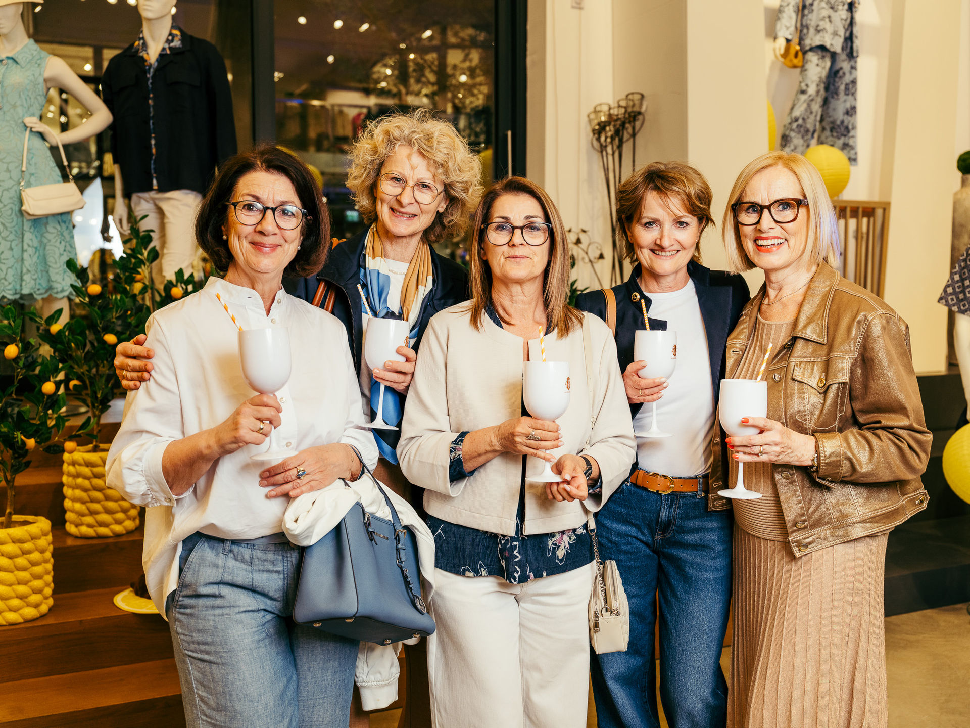 Fünf ältere Frauen mit Brillen halten weiße Gläser mit Strohhalmen und lächeln in einem Geschäft mit Modepuppen im Hintergrund.