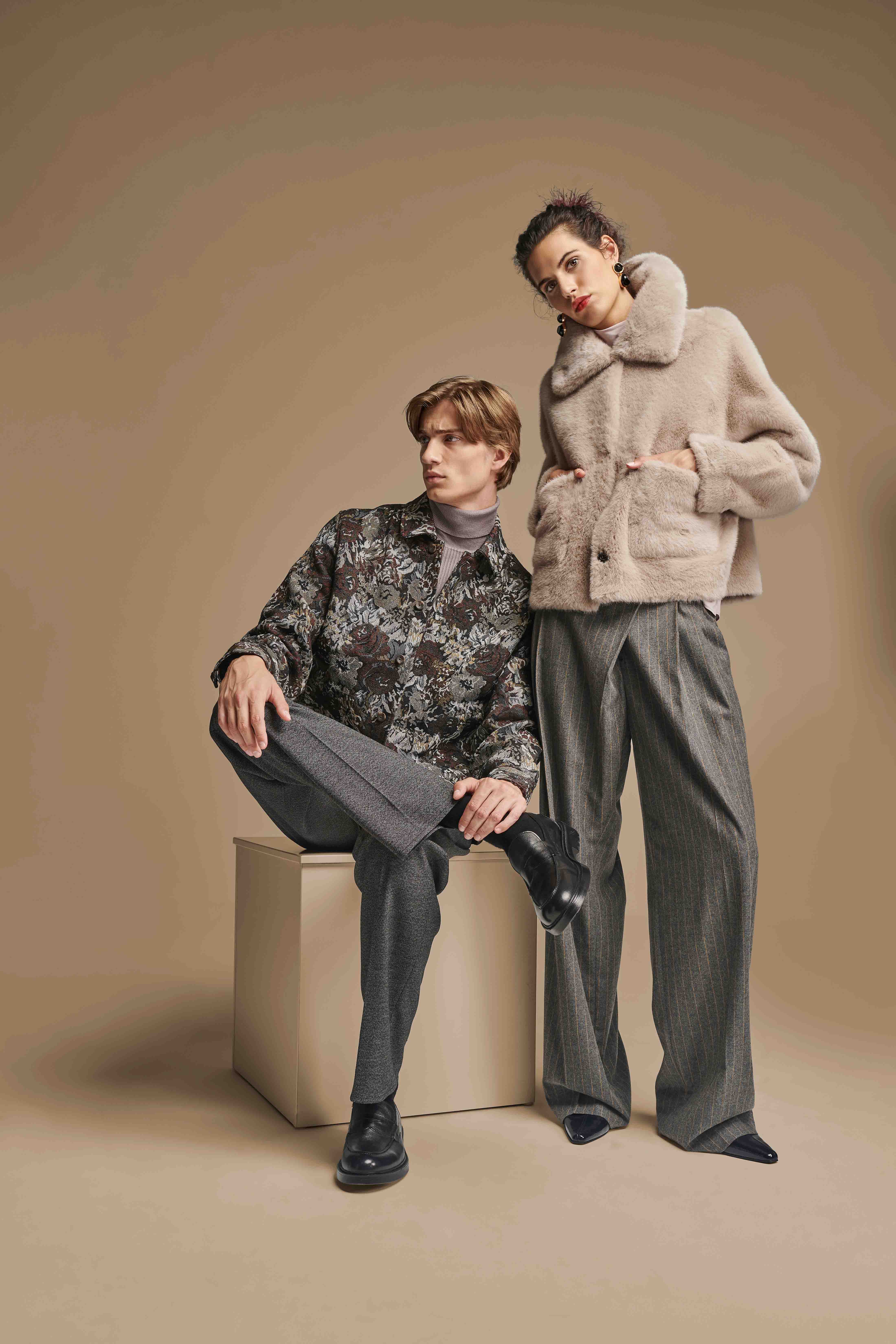 Ein sitzender Mann trägt eine dunkle Hemdjacke mit floralem Muster, graue Hose und schwarze Schuhe; eine stehende Frau neben ihm trägt eine beige Pelzjacke, graue weite Hose und schwarze spitze Schuhe vor einem einfarbigen beigen Hintergrund.