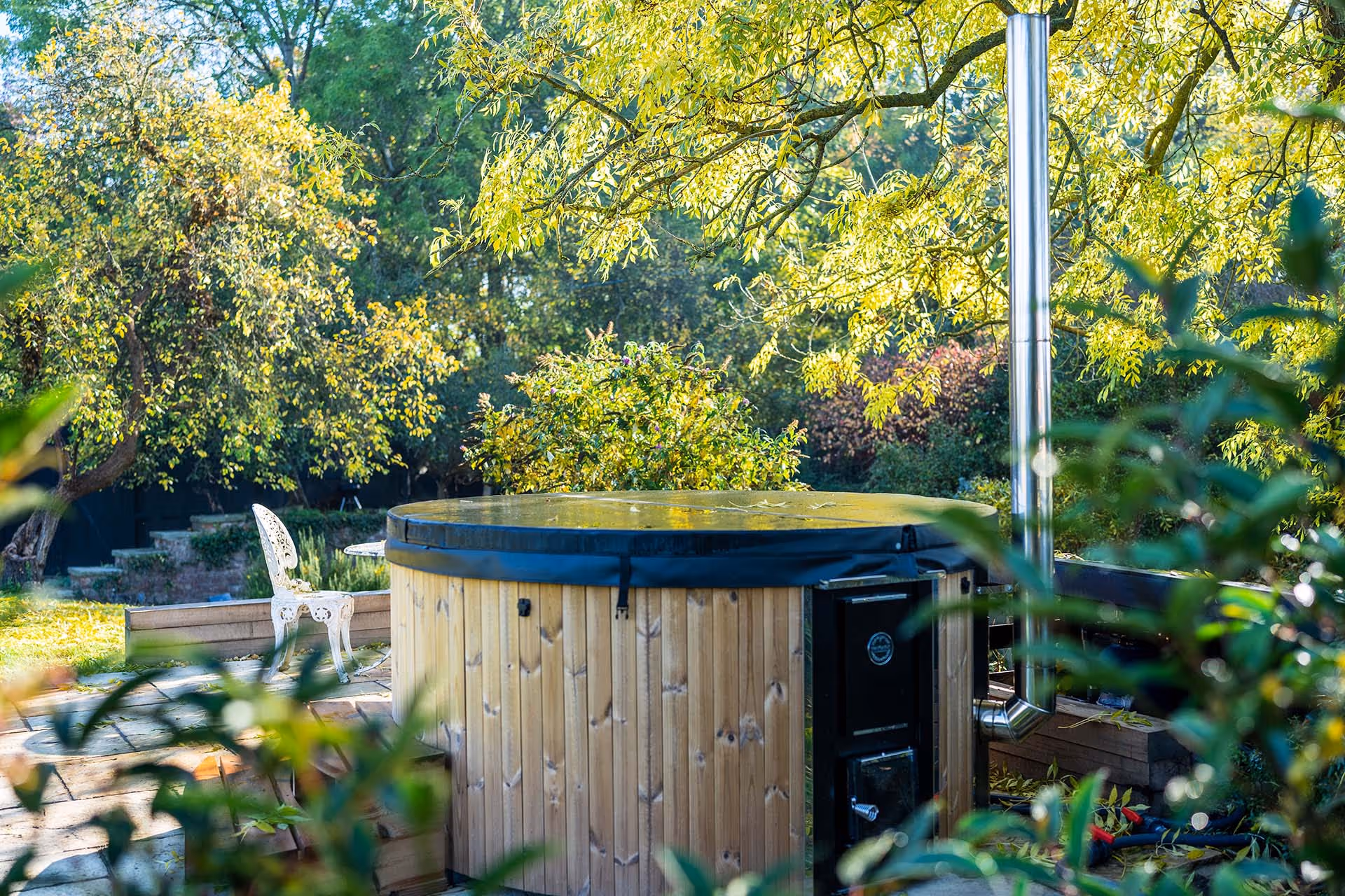 Wooden hot tub in lush garden with yellow-green trees and white chair
