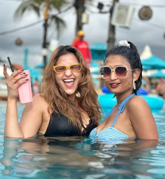 Top-down view of a woman in a yellow swimsuit floating on her back in a rectangular pool surrounded by greenery and wooden decking with cushions and drinks nearby.