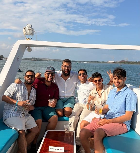 Group of six young men sitting closely together on a boat with a calm sea and partly cloudy sky in the background, all smiling and enjoying drinks on a Triptease group trip