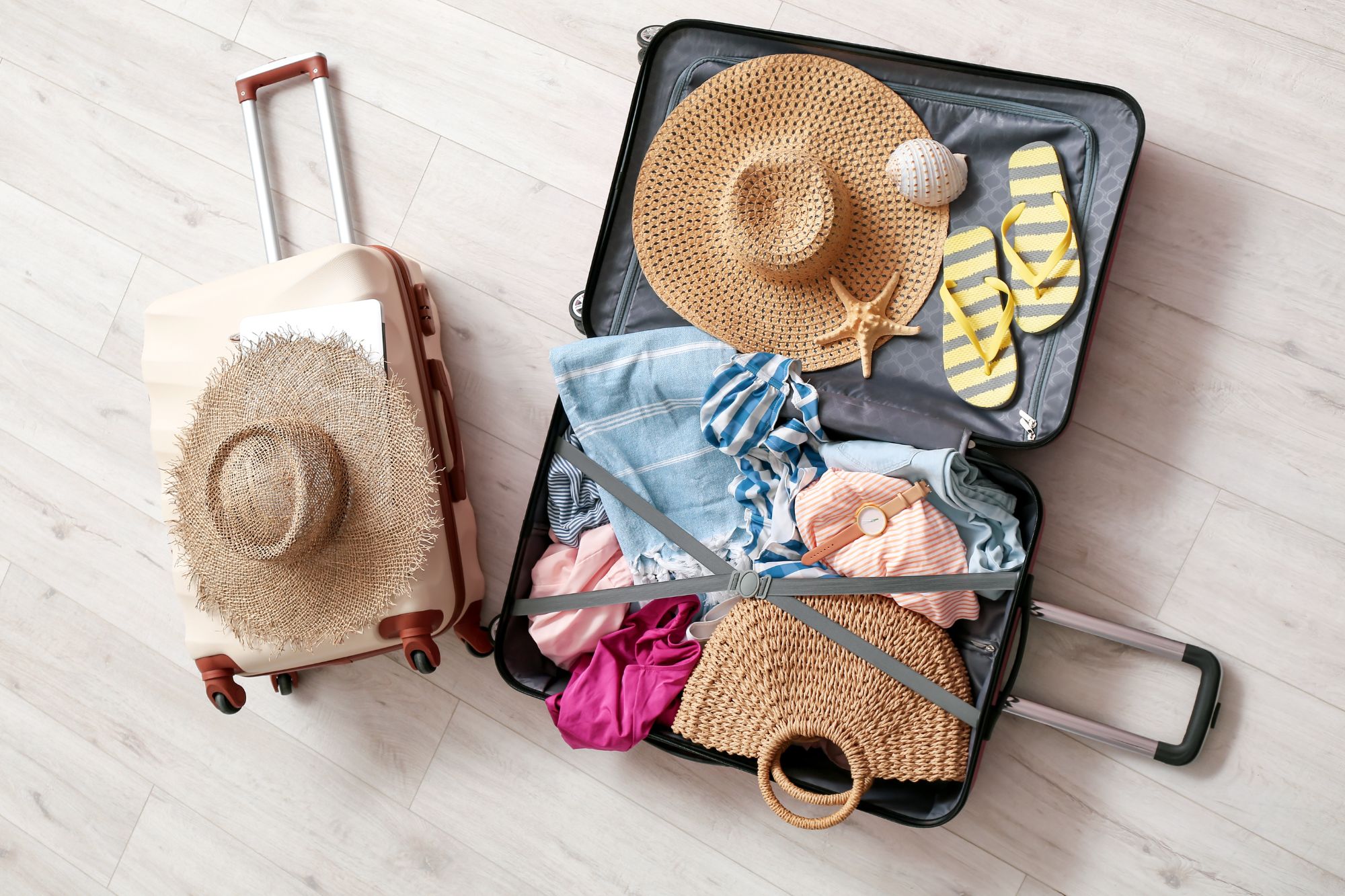 Top view of open suitcase packed with summer travel essentials including straw hats, flip-flops, striped clothes, and beach accessories.