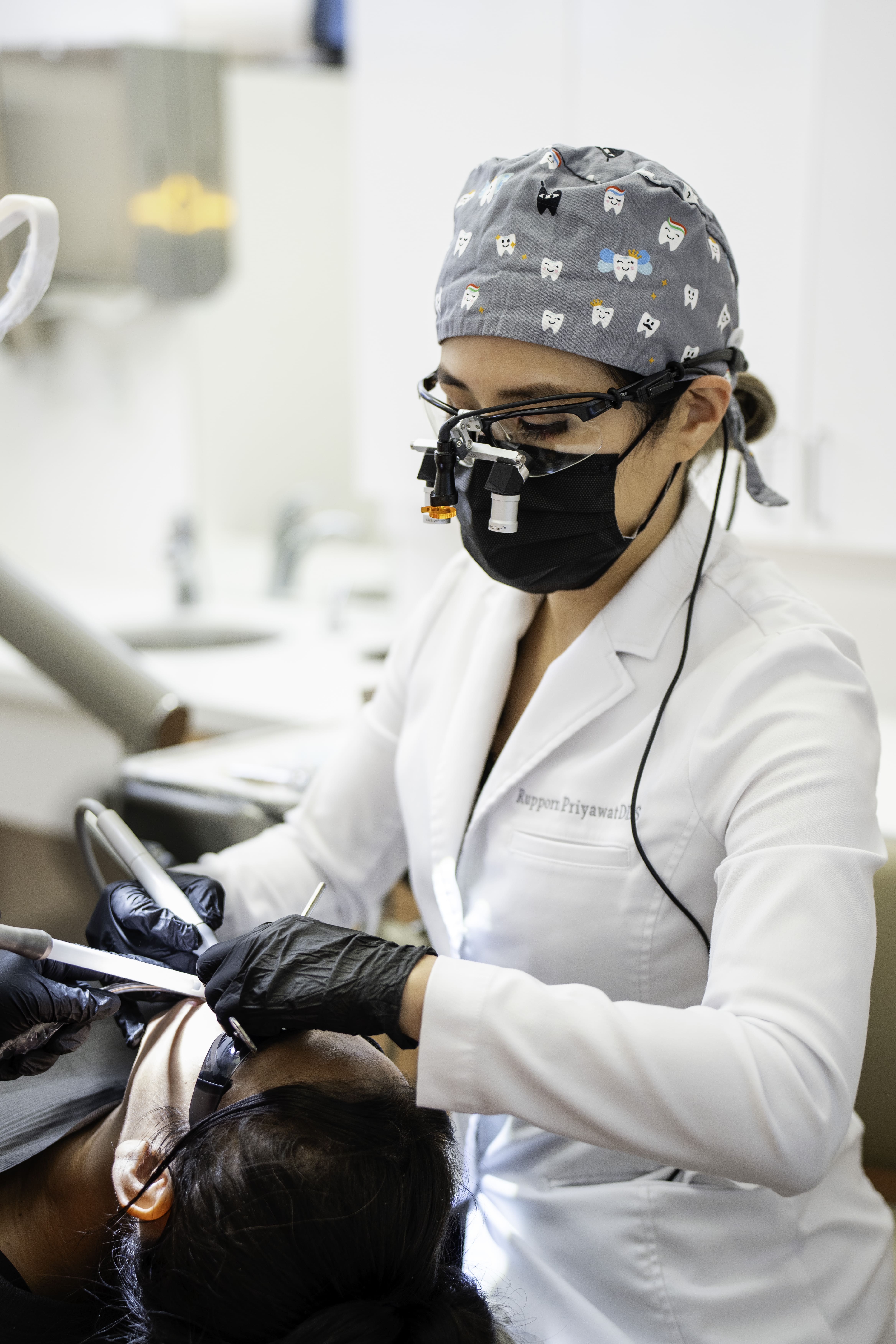 Dr. Rupporn Priyawat wearing magnifying glasses and a cap with tooth illustrations performing dental work on a patient Happy patients just got treatment at at Omnia Dental Care in Chino, CA 