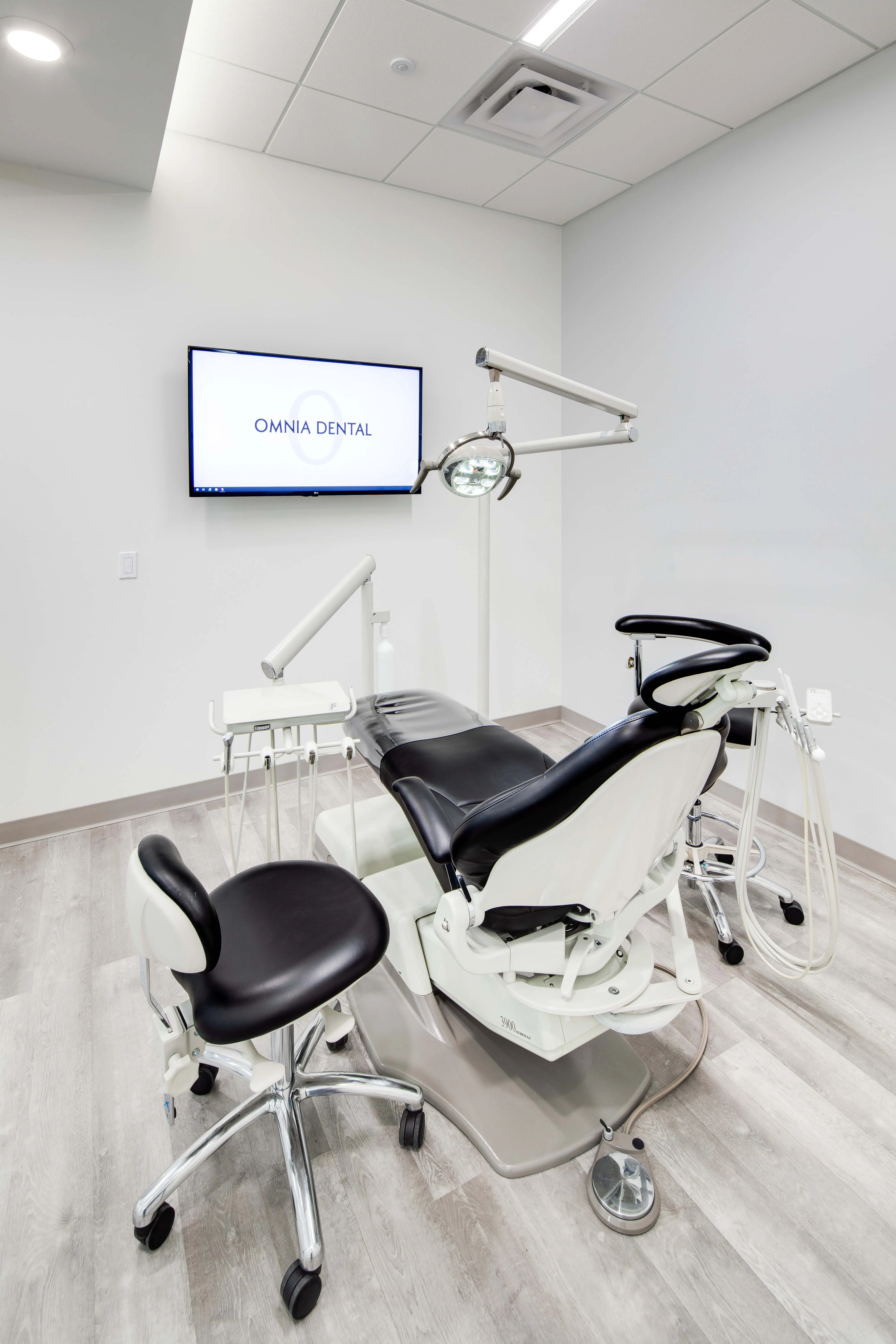 Modern dental examination room with black and white dental chair, assistant stool, overhead light, and monitor displaying 'OMNIA DENTAL'.