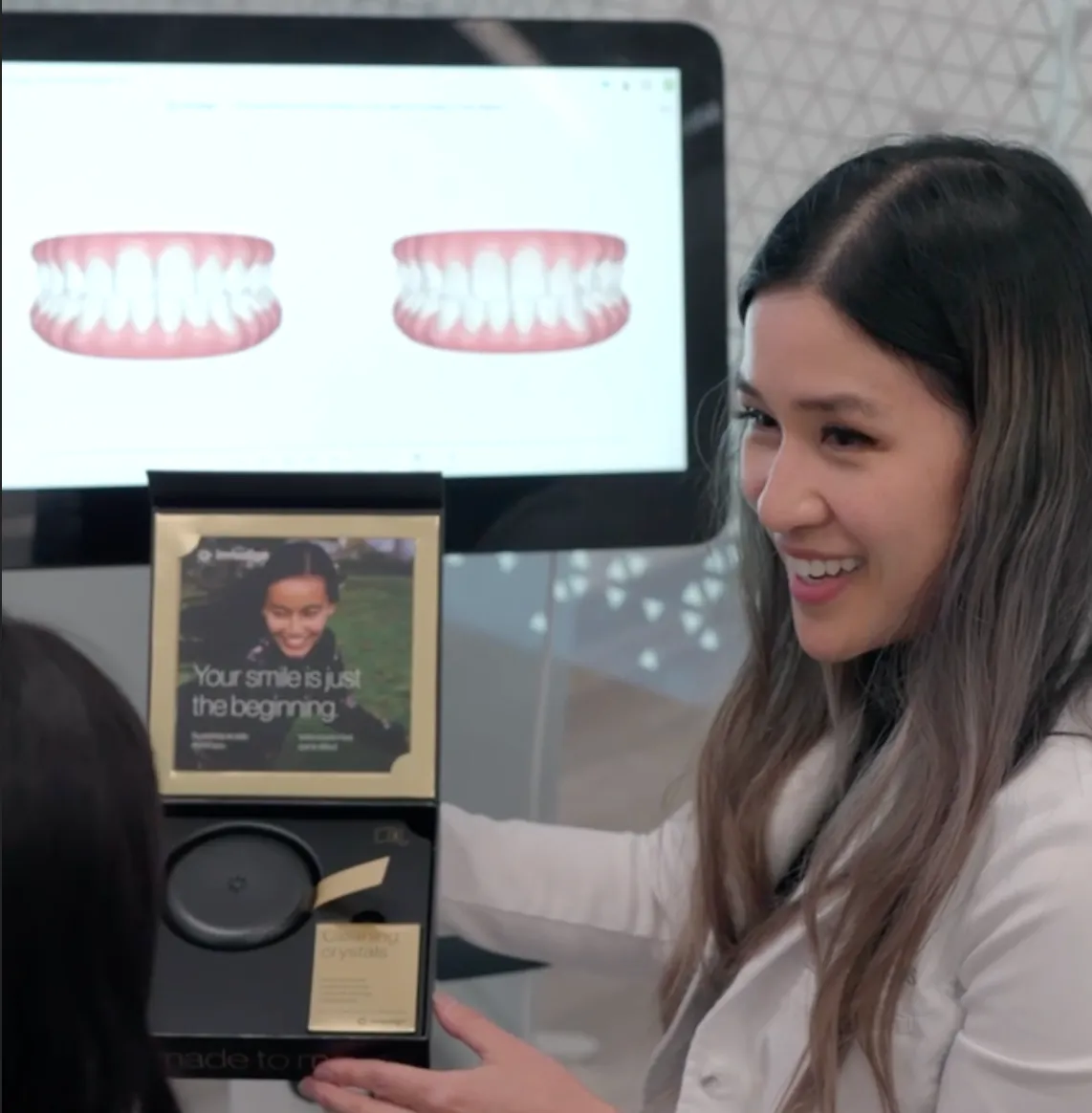 Dr. Rupporn Priyawat woman showing an open box with a dental aligner case inside, with a computer screen behind displaying two digital images of teeth at Omnia Dental Care in Chino, CA 