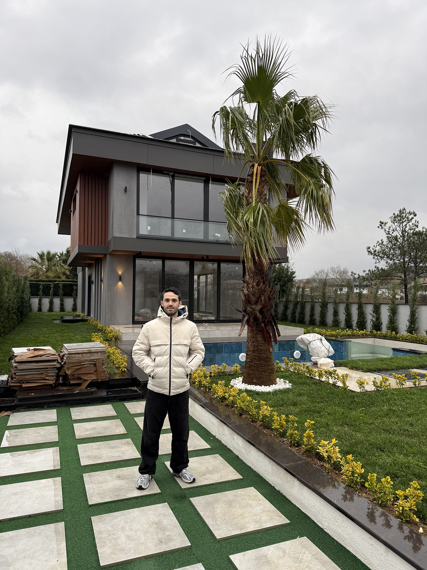 Modern white duplex with flat roofs, solar panels, glass balconies, and geometric design, surrounded by a black gate and shrubs, under clear blue sky in sunny residential neighborhood.