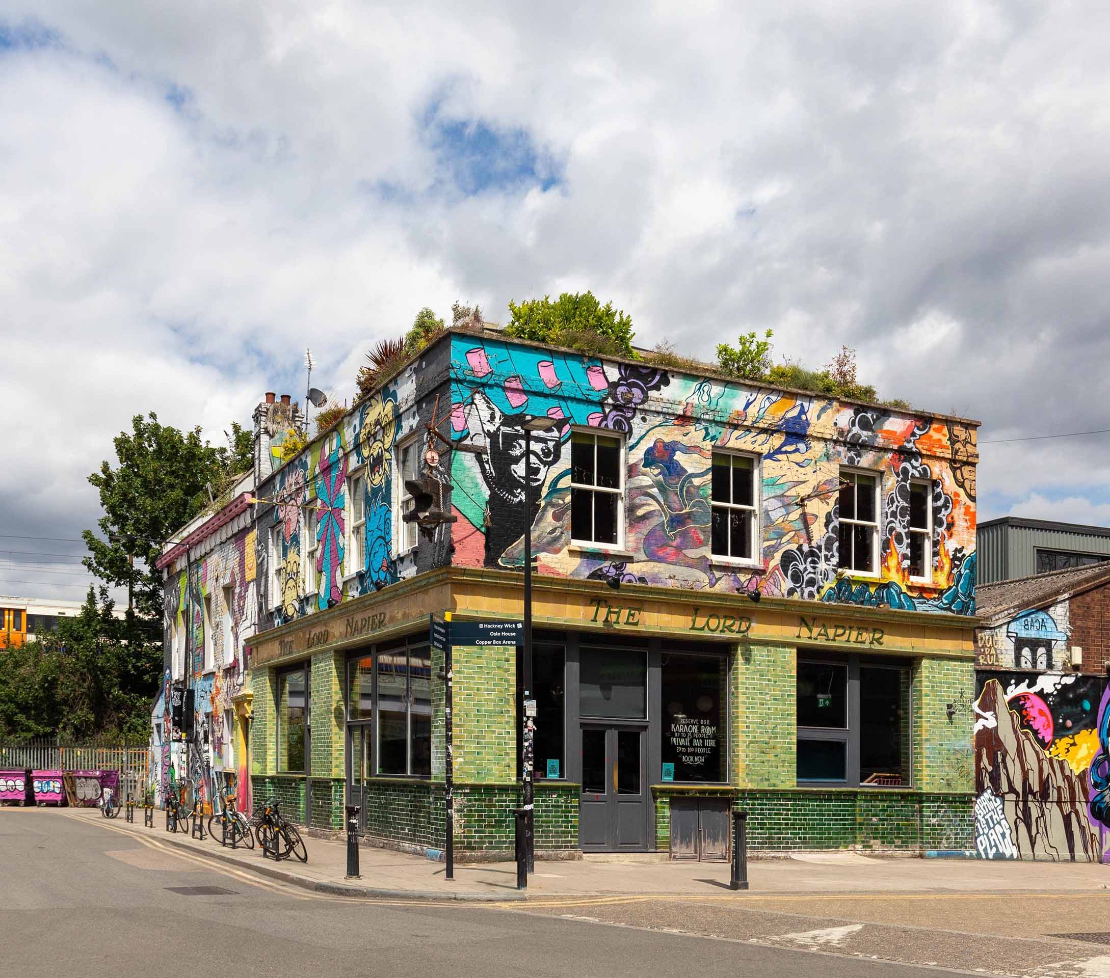 Two-story corner building with green tiles and colorful graffiti murals covering the upper walls, named The Lord Napier pub with bicycles parked outside.
