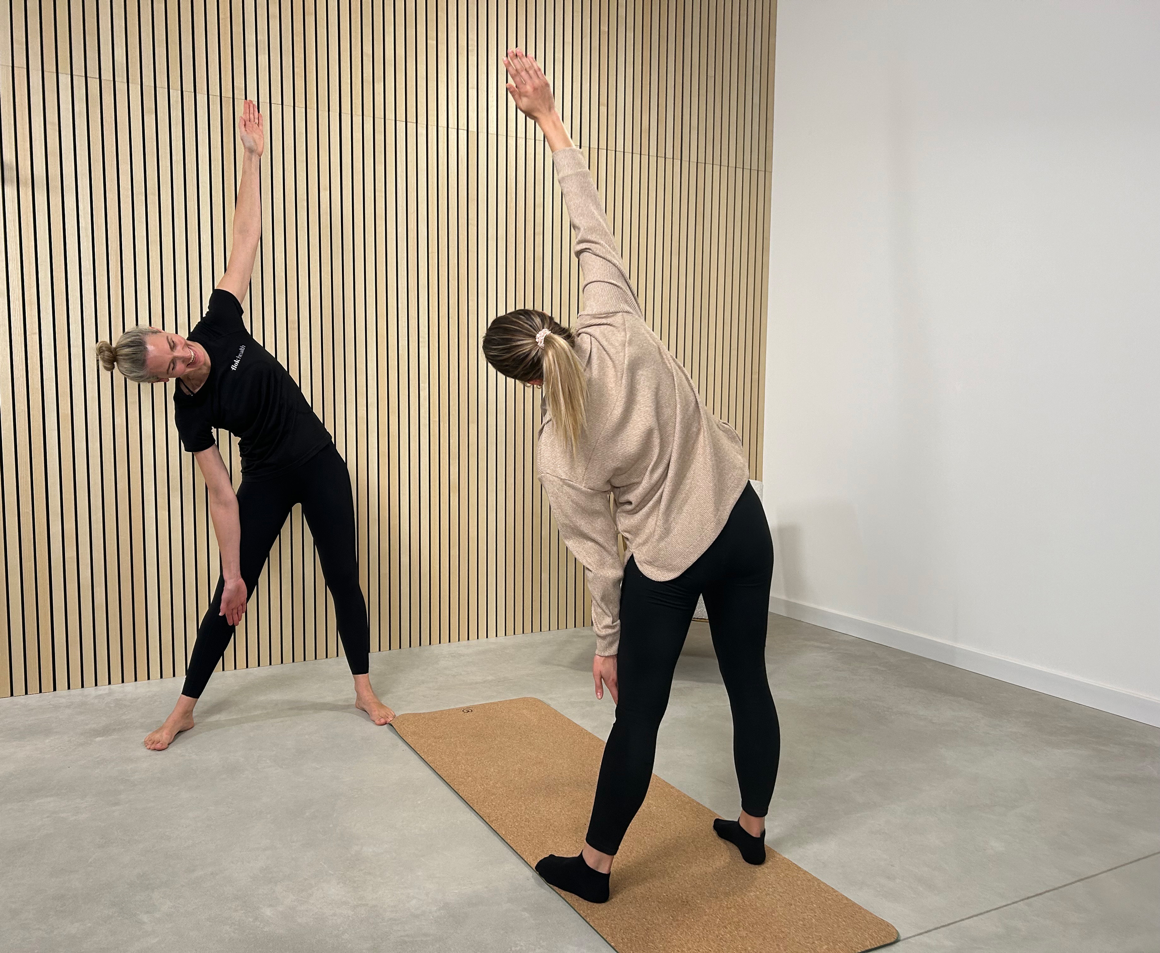 Two women practicing side stretches indoors on a yoga mat, one wearing black workout clothes and the other in a beige sweater and black leggings.