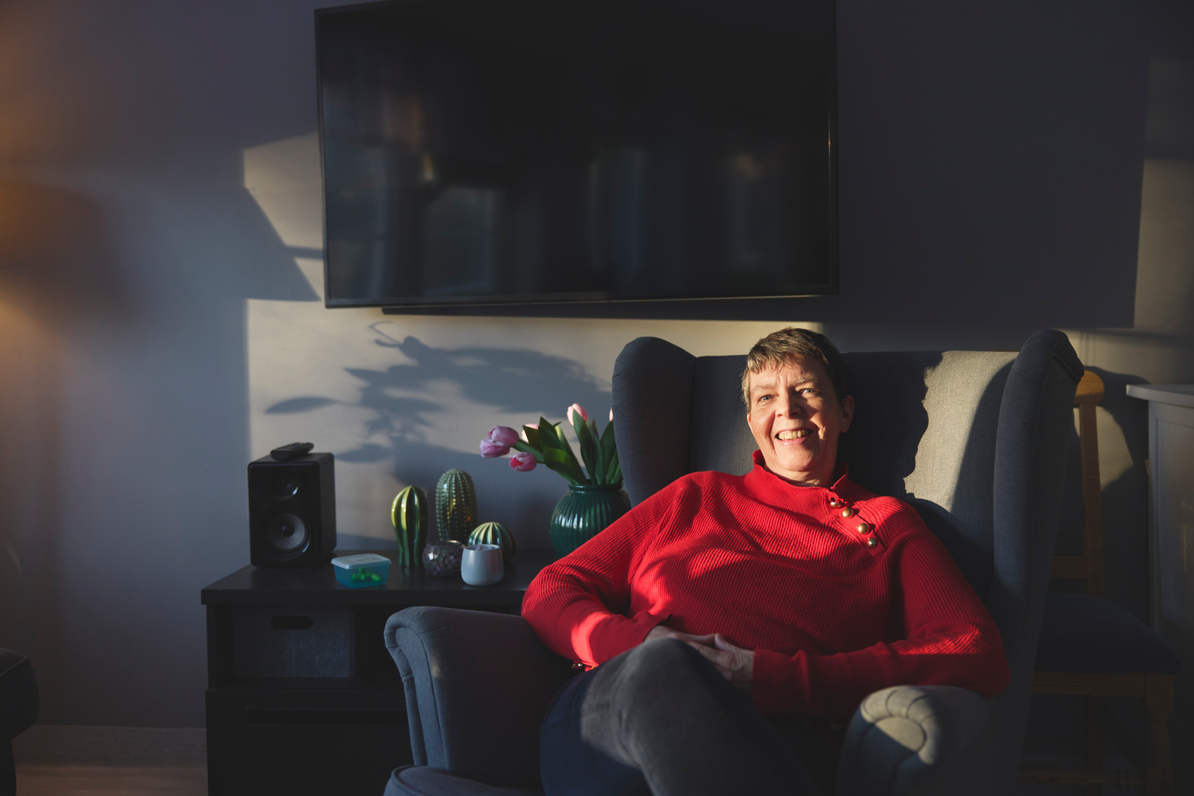Smiling woman in a red sweater sitting on a gray armchair in a dimly lit living room with a TV and flowers in the background.