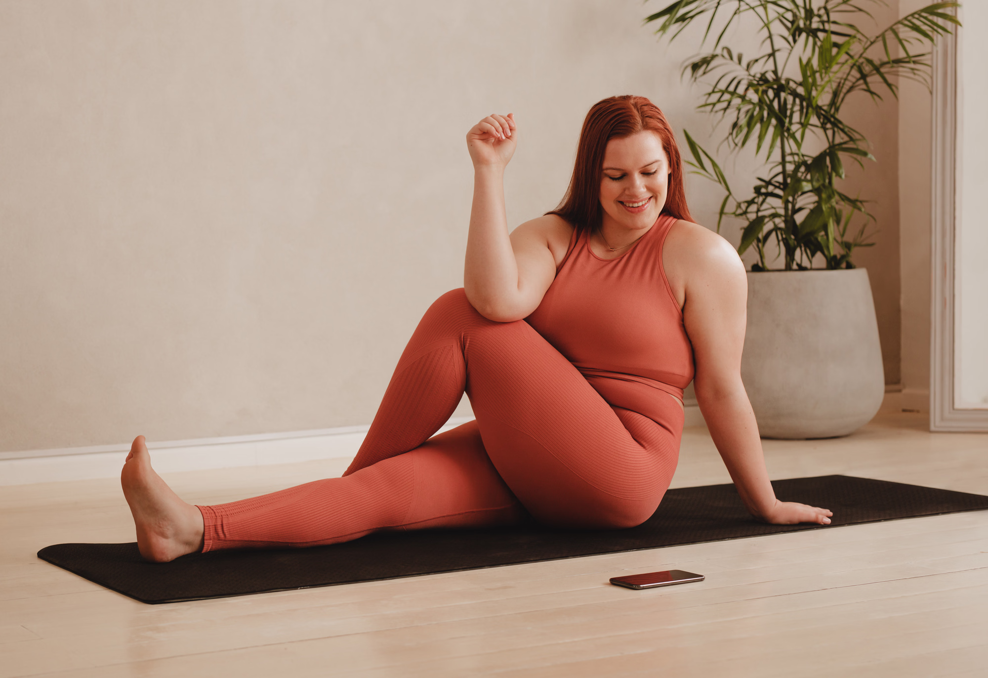 Smiling woman in rust-colored workout clothes sitting on a black yoga mat performing a seated spinal twist stretch.