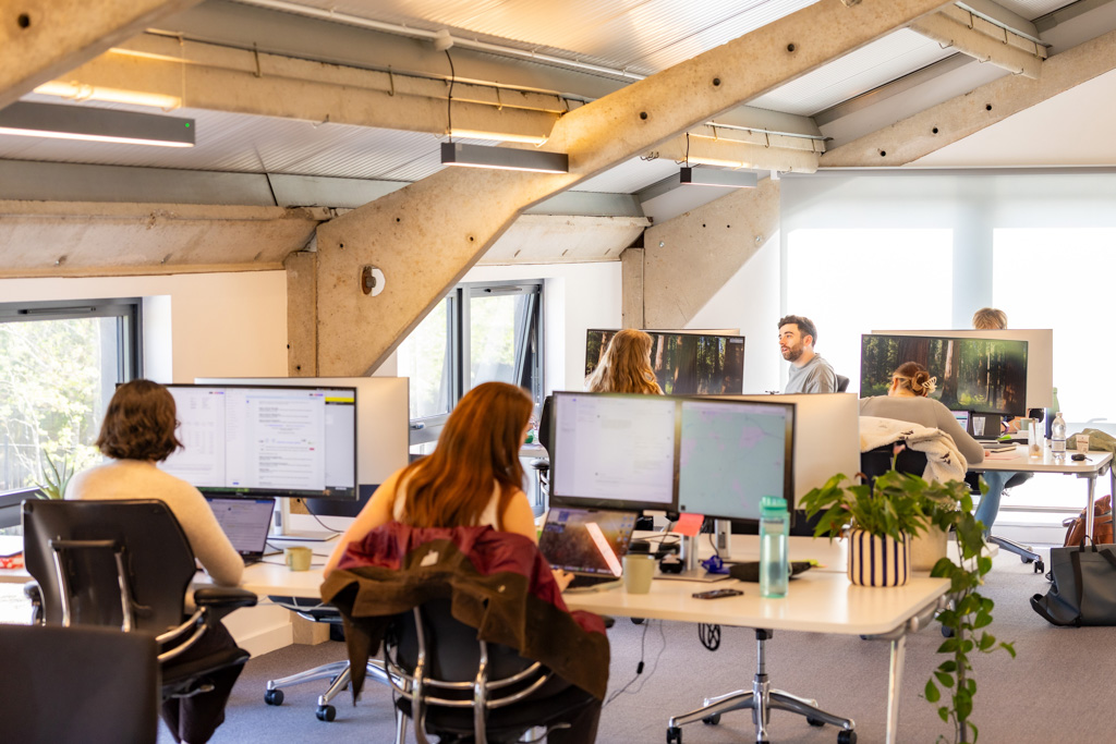 Modern open-plan office with people working at desks with multiple monitors and natural light coming through large windows.