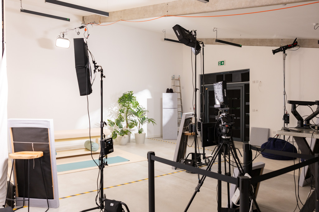 Interior of a video studio with lights, camera equipment, potted plants, and a yoga mat on the floor.