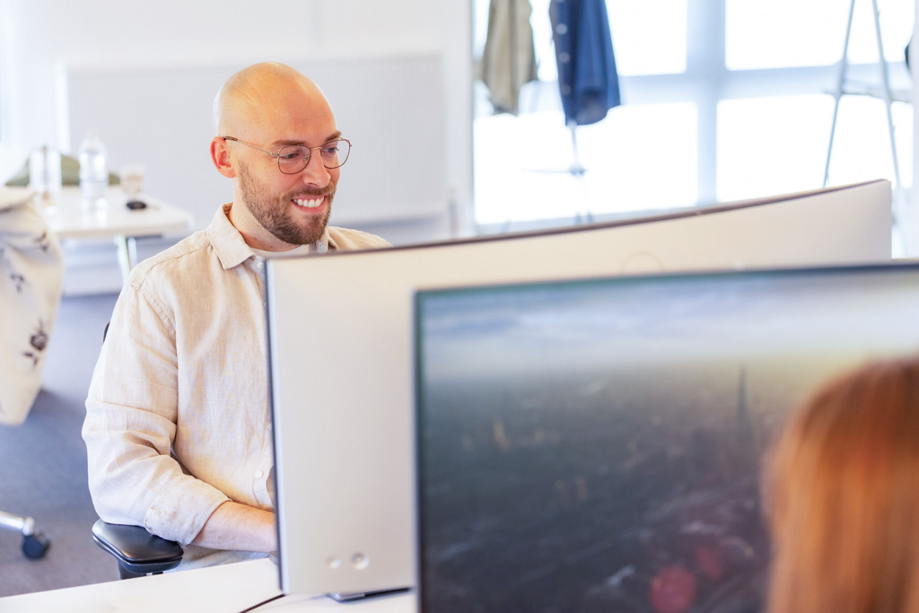 Smiling bald man with glasses sitting at a desk in front of dual computer monitors in a bright office.