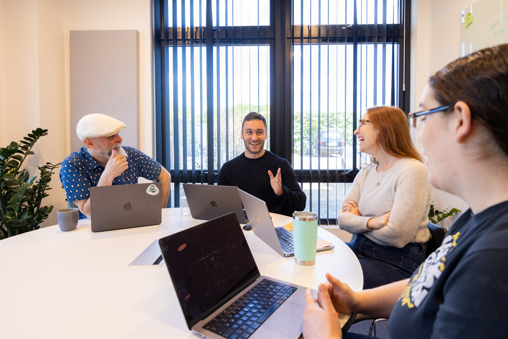 Four people sitting around a white table in an office having a lively discussion with laptops in front of them.