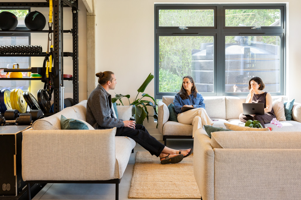 Three people sitting on beige sofas in a bright room next to an indoor gym.