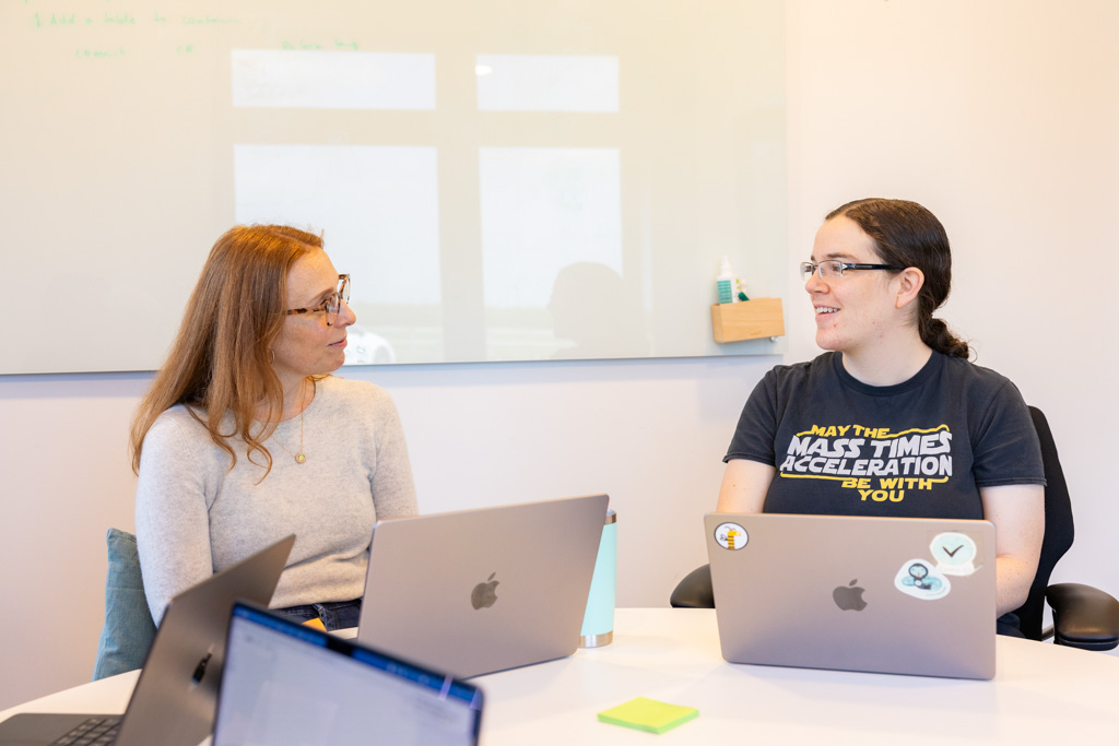 Two women sitting at a table with laptops, engaged in conversation in a meeting room with a whiteboard behind them.