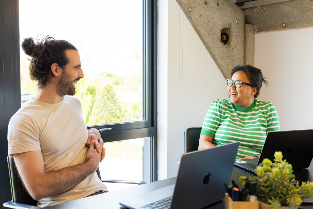 Two colleagues smiling and chatting at a table with laptops near a large window in a bright office.