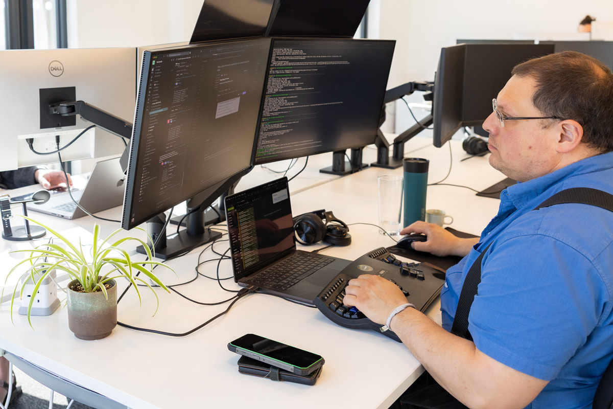 Man in blue shirt working at a desk with dual monitors, a laptop, ergonomic keyboard, and smartphone.