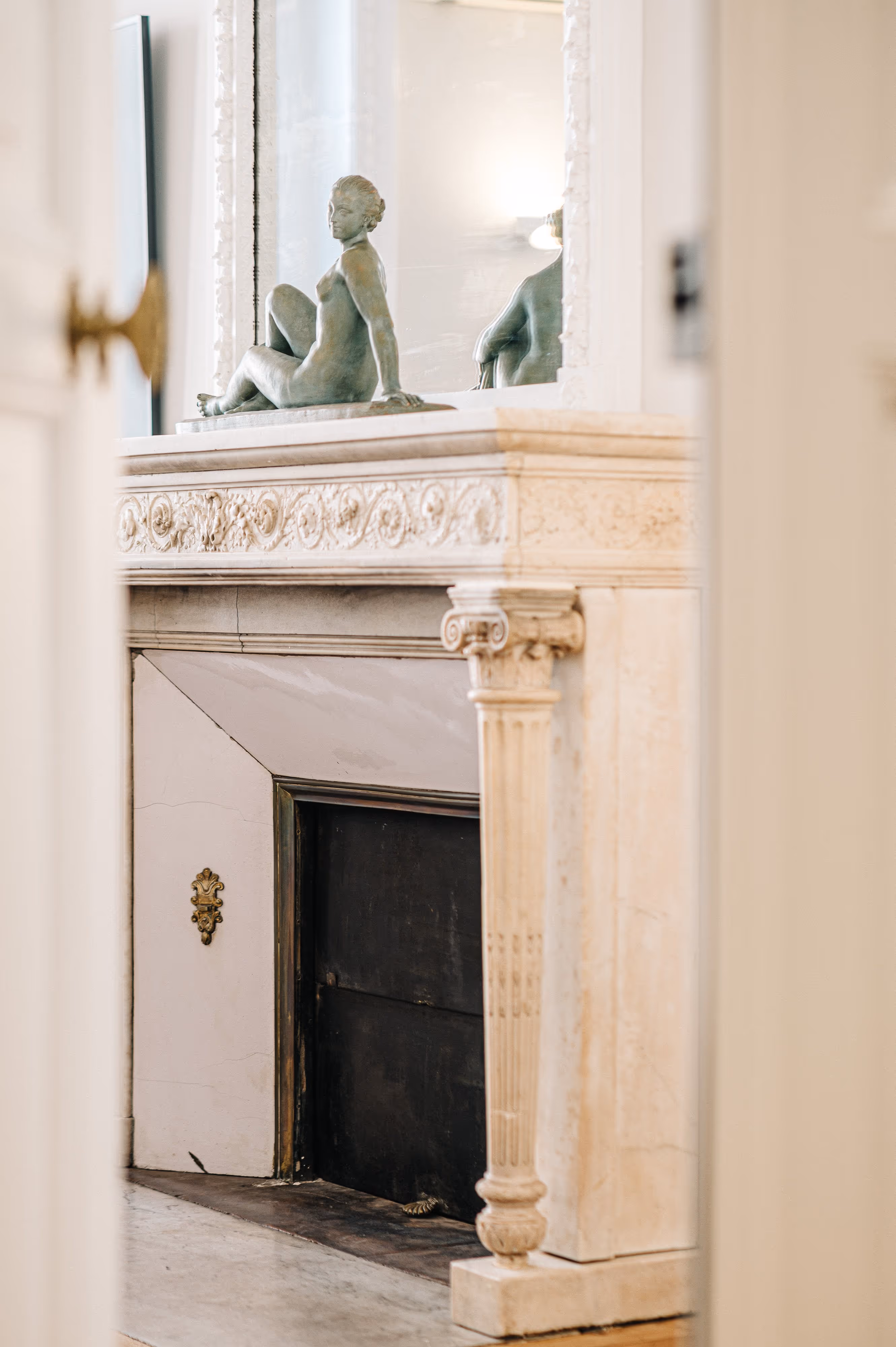 Vue à travers une porte entrouverte sur une cheminée ancienne ornée d'une statue d'une femme assise devant un grand miroir.
