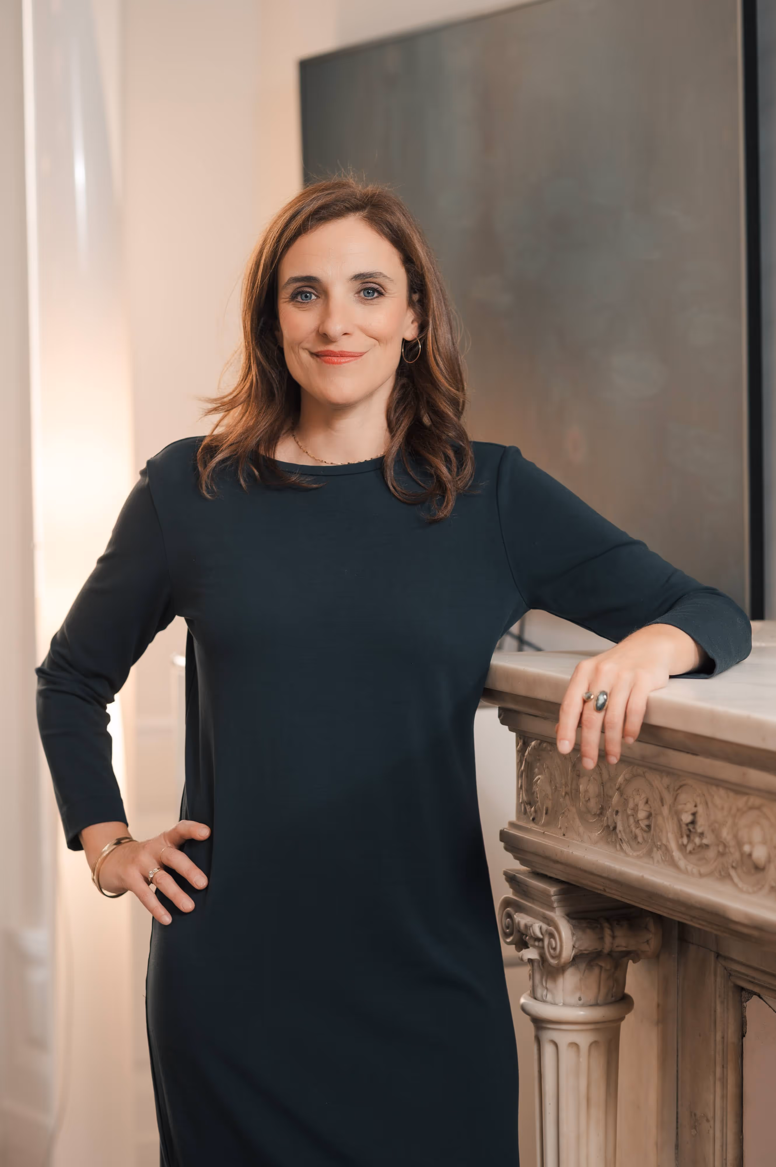 Femme souriante en robe noire posant près d'une cheminée en marbre sculpté.