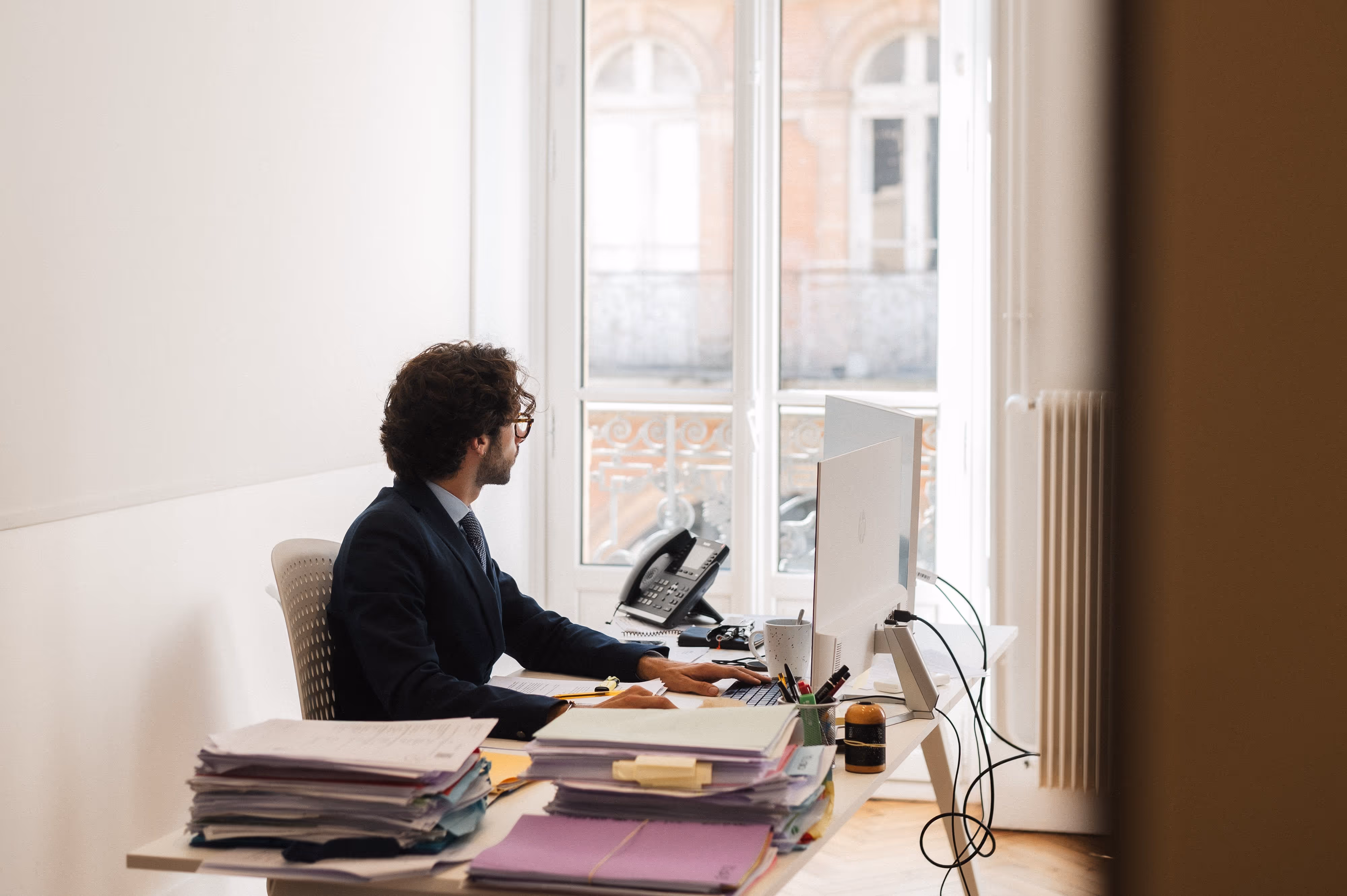 Homme en costume assis à un bureau encombré de piles de dossiers, travaillant sur un ordinateur avec un téléphone de bureau à côté.