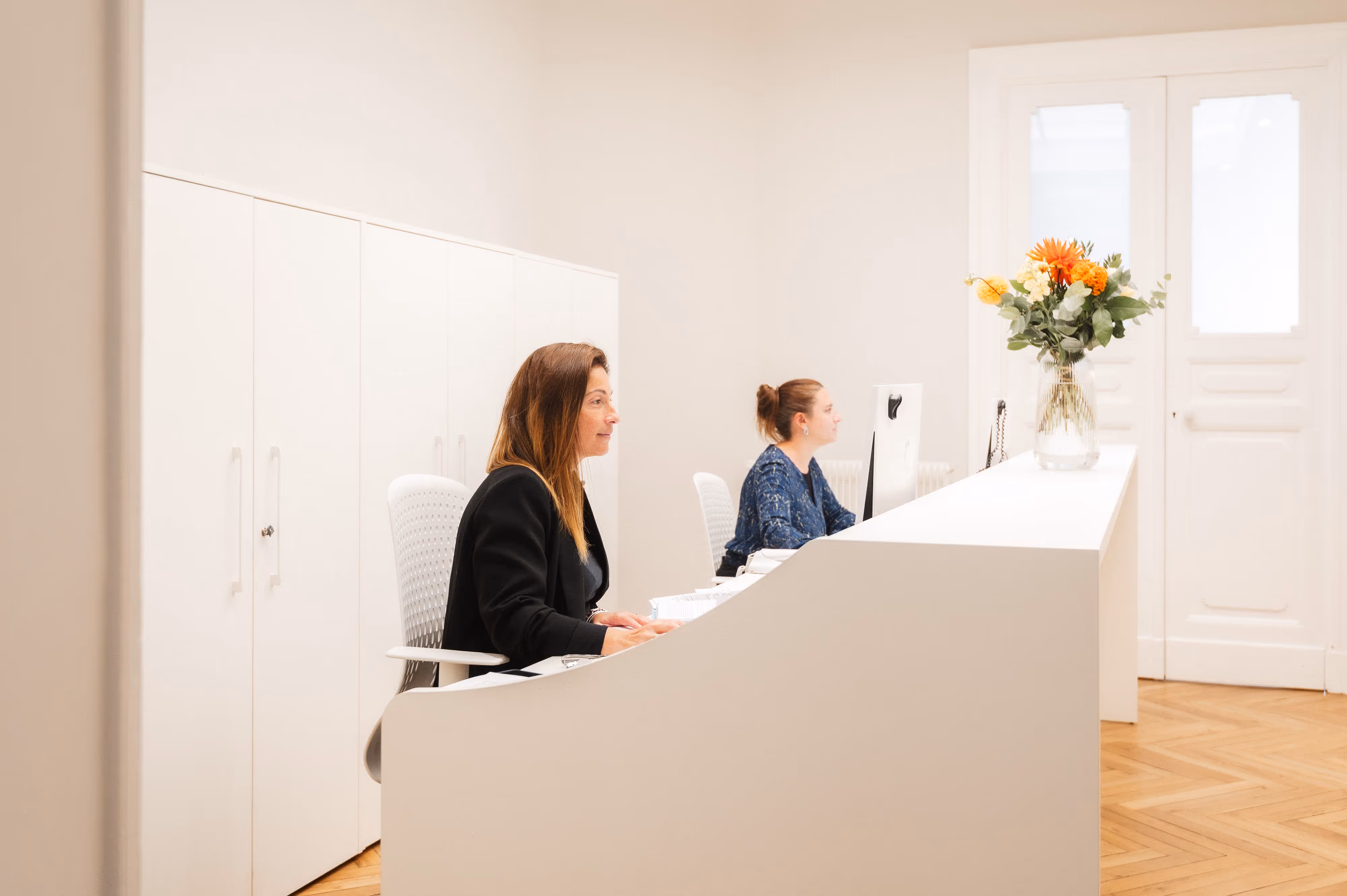 Deux femmes assises à un bureau blanc dans un bureau clair avec un vase de fleurs colorées sur le comptoir.