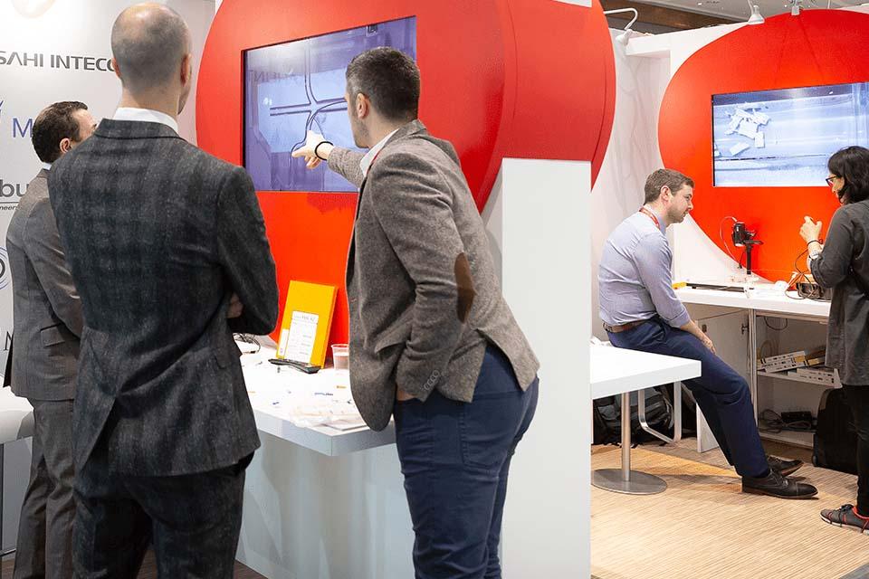 Group of professionals in business attire interacting and discussing two screens displaying technical or security footage in a modern office or exhibition space.