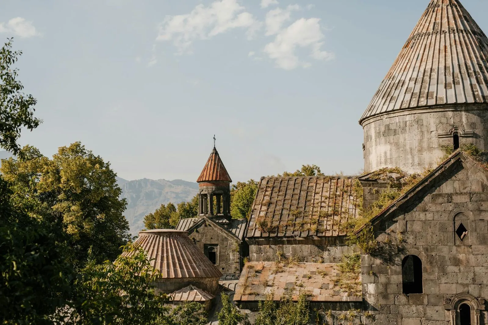 Historic Armenian monastery with stone domes and rustic rooftops, surrounded by lush green trees and mountains.