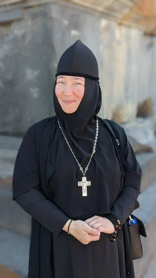 Smiling Armenian nun in black robes and a cross necklace, standing outside a historic monastery.