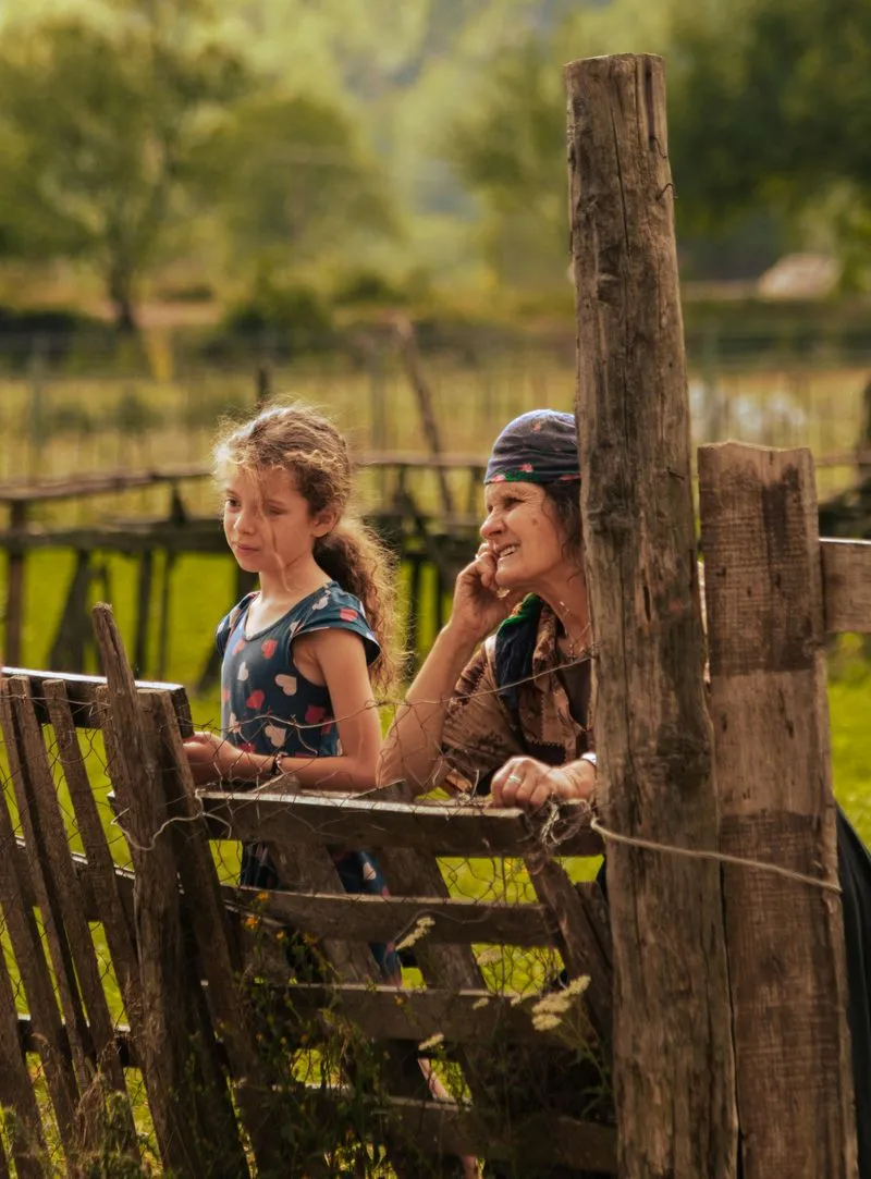 An elderly woman in a headscarf and a young girl standing by a rustic wooden fence in an Albanian countryside village.
